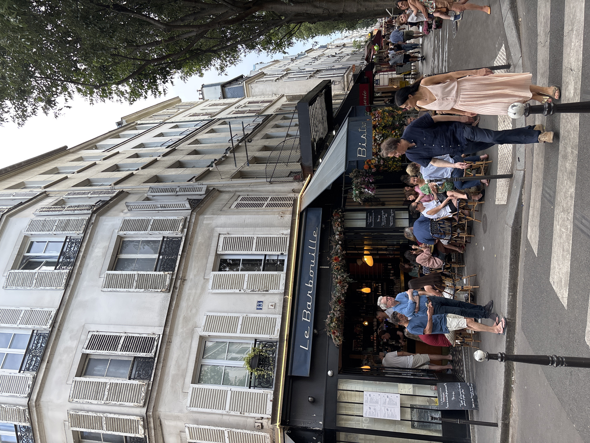 This is a street-level, eye-level, medium shot of a Parisian street scene. The buildings are typical Haussmannian architecture, with pale grey facades, white shutters, and ornate iron balconies. The image captures the exterior of a restaurant and bistro, "Le Barbouille," with outdoor seating. Several people are visible, some seated at tables and others walking on the street and sidewalk.
In the foreground, two individuals are walking across a crosswalk. A man in a dark blue button-down shirt and dark pants is on the left, and a woman in a light pink pleated dress is on the right. Behind them, on the sidewalk, a man in a blue button-down shirt and khaki shorts stands with his arms crossed, looking to his right. To his left, another man in a blue t-shirt and khaki shorts is looking down at his phone.
The restaurant "Le Barbouille" has a black awning with its name in white script. Floral decorations hang above the entrance. Inside the restaurant, patrons are seated at tables and standing at the bar. Outside, tables and chairs are arranged along the sidewalk, with diners enjoying their meals. Further down the street, other businesses with awnings and outdoor seating are visible, and more pedestrians are walking. The overall atmosphere suggests a lively, urban setting in Paris, likely during the daytime. The trees lining the street indicate a pleasant urban environment.