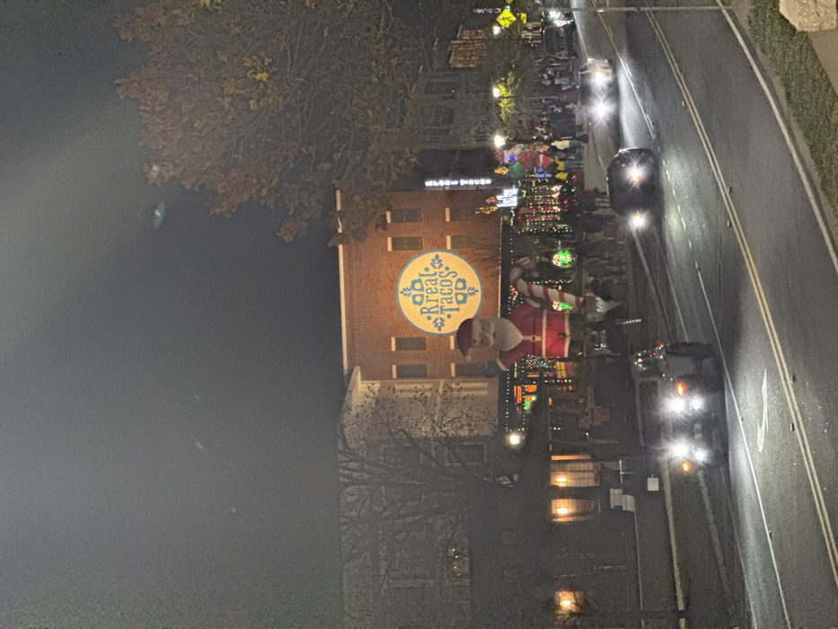 A festive night scene unfolds with "Kreal Tacos" displayed prominently on a brick building. An inflatable Santa and candy cane stand alongside colorful holiday lights lining the street. Cars with headlights on drive along the adjacent road. Silhouetted figures gather on the sidewalk, suggesting a lively event. Trees flank the scene, adding depth to the nighttime setting.