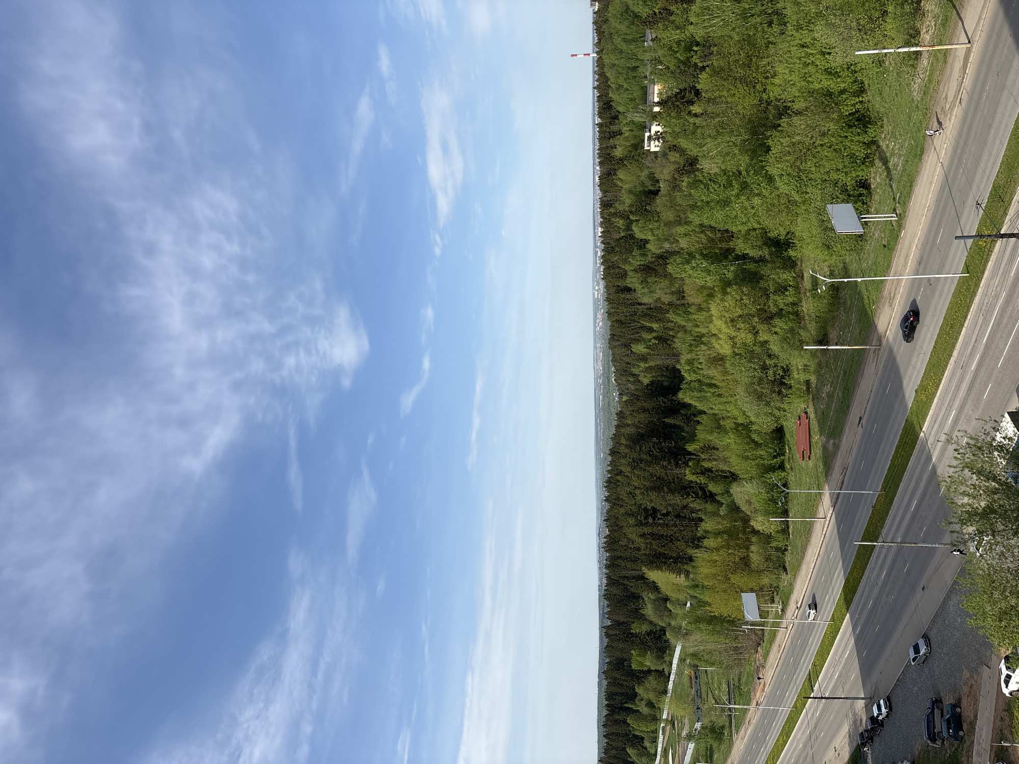 The image presents an elevated view showing a clear division between a blue sky with scattered clouds on the left and a dense forest and urban road on the right. The road runs parallel to the forest, featuring multiple lanes of traffic and a line of streetlights. Several cars are visible on the road and parked along the side. In the distance, within the forest area, there are some buildings, including a tall industrial chimney with a red and white striped top. The location is Izhevsk, Russia.