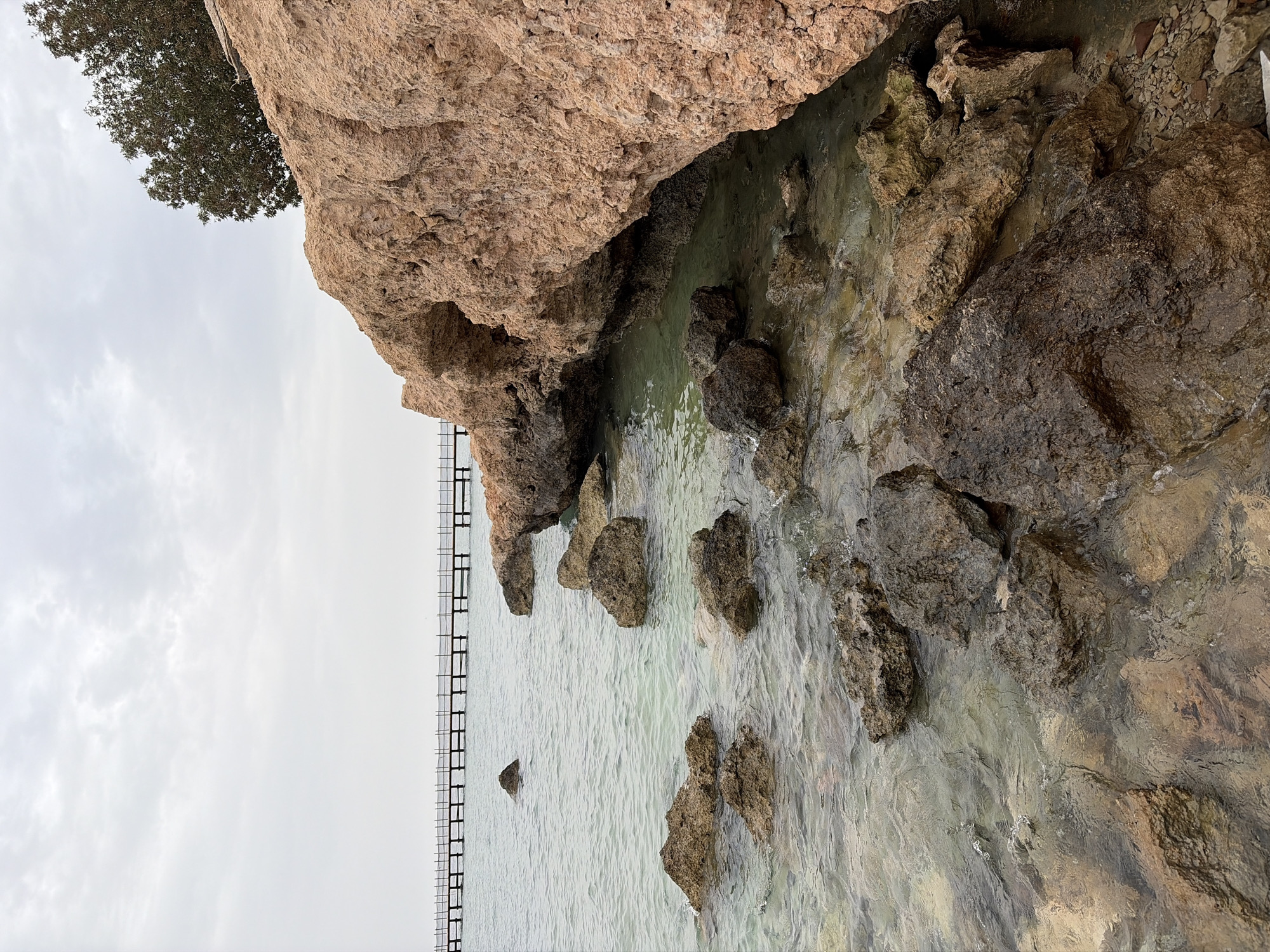 The image shows a rocky coastal scene in Sharm El Sheikh, Egypt. A large, light brown rock formation dominates the upper portion of the frame. Beneath it, clear water laps against a shoreline comprised of smaller rocks. A metal pier or walkway extends into the water on the left side of the frame, suggesting human intervention in the landscape. The sky is overcast, with a grey, cloudy expanse above. In the upper left corner, a tree is visible. The water is relatively clear, revealing submerged rocks and varying shades of green and brown.
