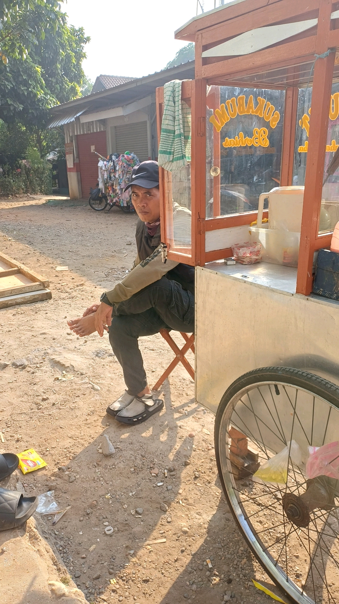 A man wearing a black baseball cap with a Nike logo, a dark jacket over a maroon scarf, and dark pants sits on a folding chair next to a food cart. The food cart has a wooden frame and glass panels displaying text that reads "SUKABUMI". Inside the cart are containers and a large pitcher. The man is looking towards the camera with his left leg crossed over his right. His right foot is bare, and he is wearing sandals on his left foot. The ground is dirt and gravel, with several cigarette butts scattered around. In the background, there is a building with a red facade and a corrugated metal roof, as well as trees and a bicycle loaded with items. A large bicycle wheel is prominently featured in the foreground on the right side of the image. The scene appears to be outdoors, likely in a street or market area in Gunung Putri, Indonesia, given the context of the food cart and the surrounding environment.