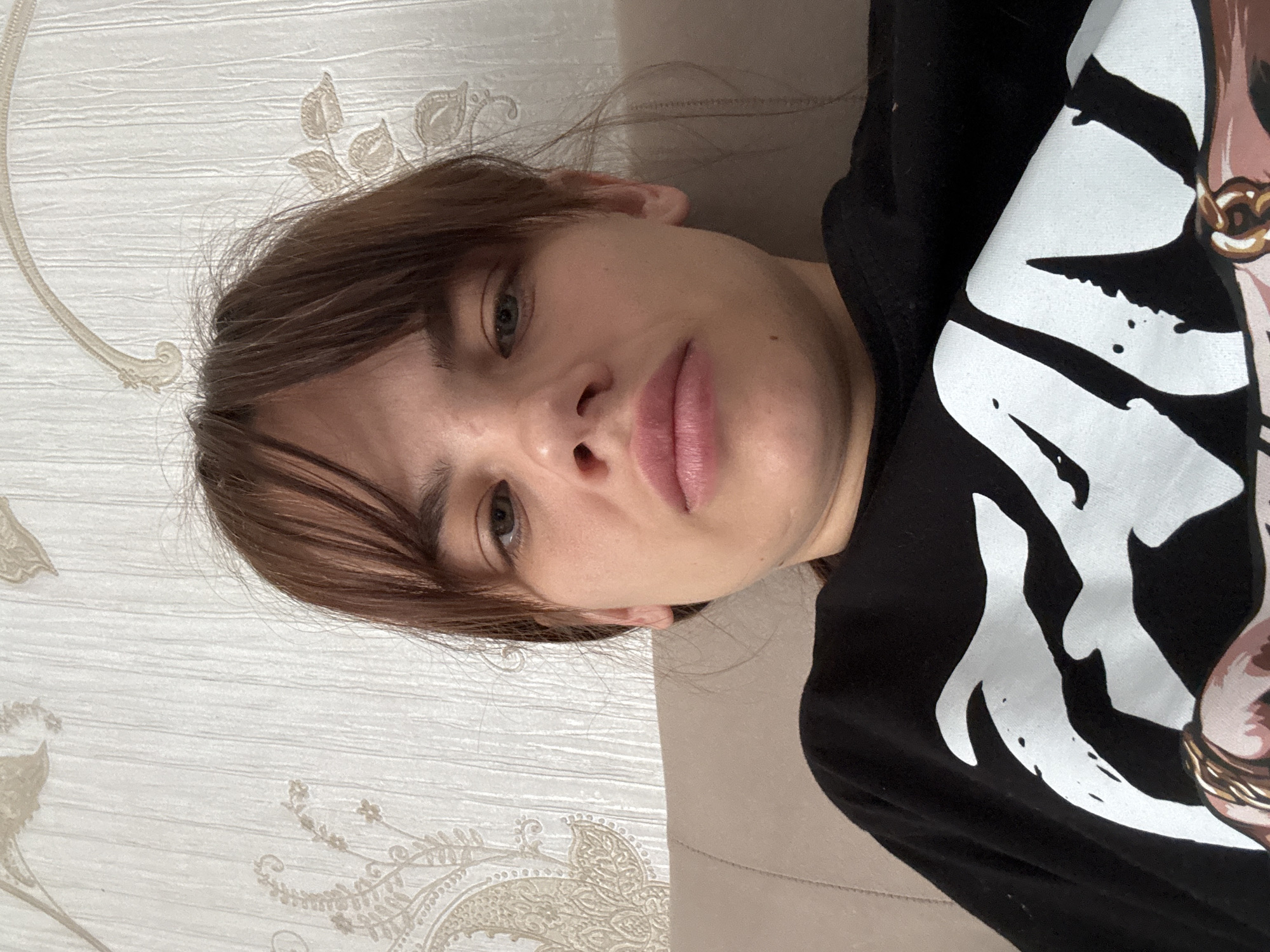 A woman with brown hair and light eyes is lying on a beige surface. She is wearing a black shirt with a white graphic design. The background is a light-colored wall with a textured floral pattern. The photo appears to have been taken indoors. There is no information to confirm this photo was taken in the city of Rostov-on-Don, Russia.