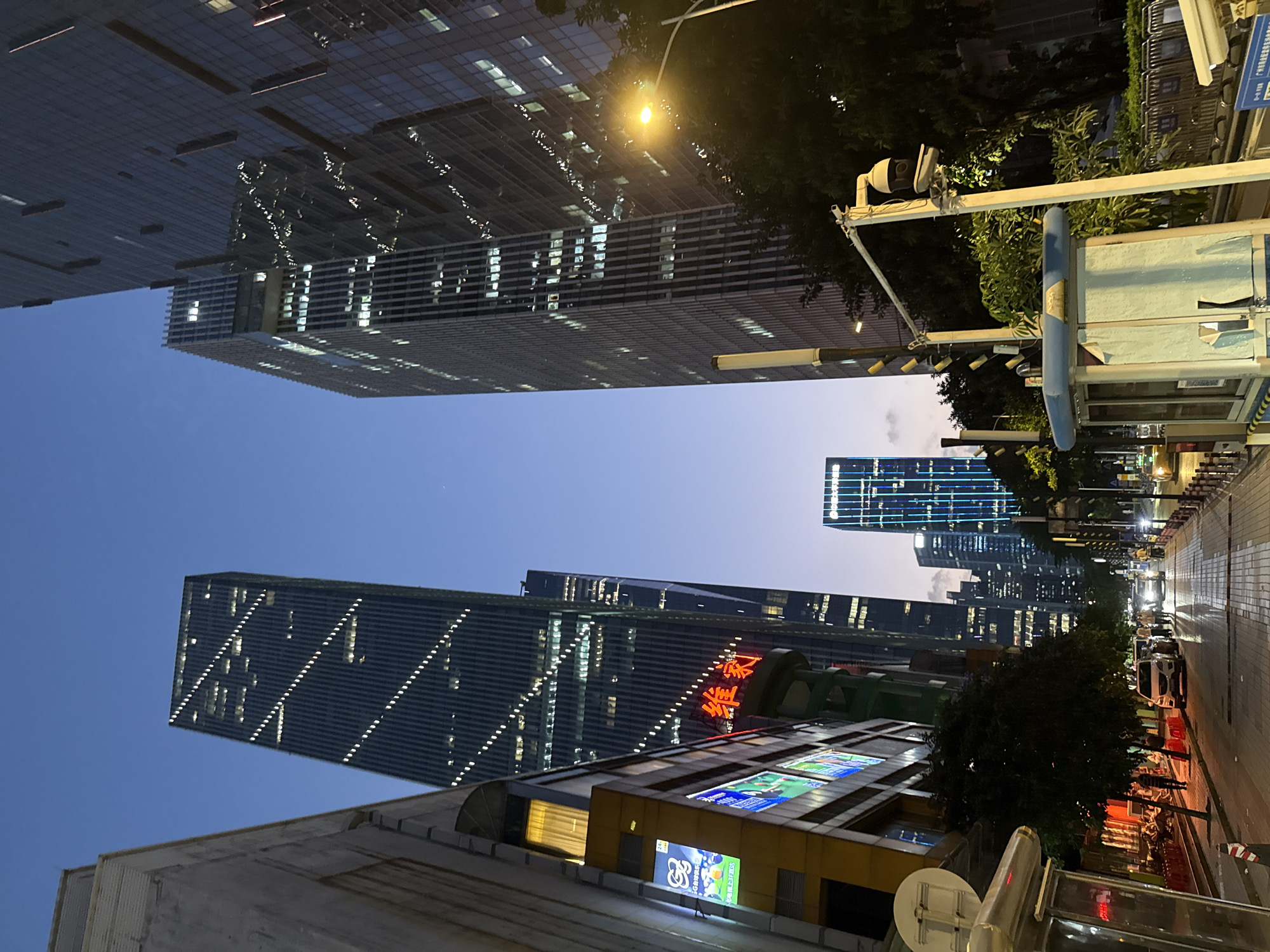 The image shows an evening street scene in the Tianhe District of Guangzhou, China. Several high-rise buildings dominate the background, illuminated with lines of lights, suggesting offices or residences. A street runs along the right side of the image, with a few parked cars. Streetlights cast a bright glow, reflecting on the wet pavement. A bus shelter and foliage are situated between the street and the base of one of the buildings. The building at the street level appears to have illuminated advertisements and signage. The sky is dusky, indicating either early evening or early morning twilight.