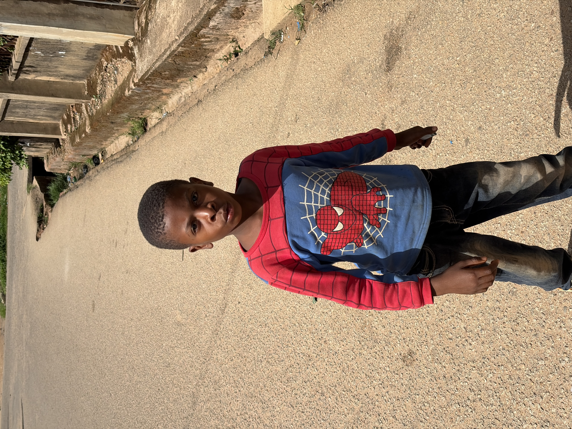 A young boy stands on a paved road in Jos, Nigeria. He is wearing a Spider-Man themed shirt and jeans. The road appears to be beside a low stone wall bordering an older building or structure. The light suggests it is a sunny day.