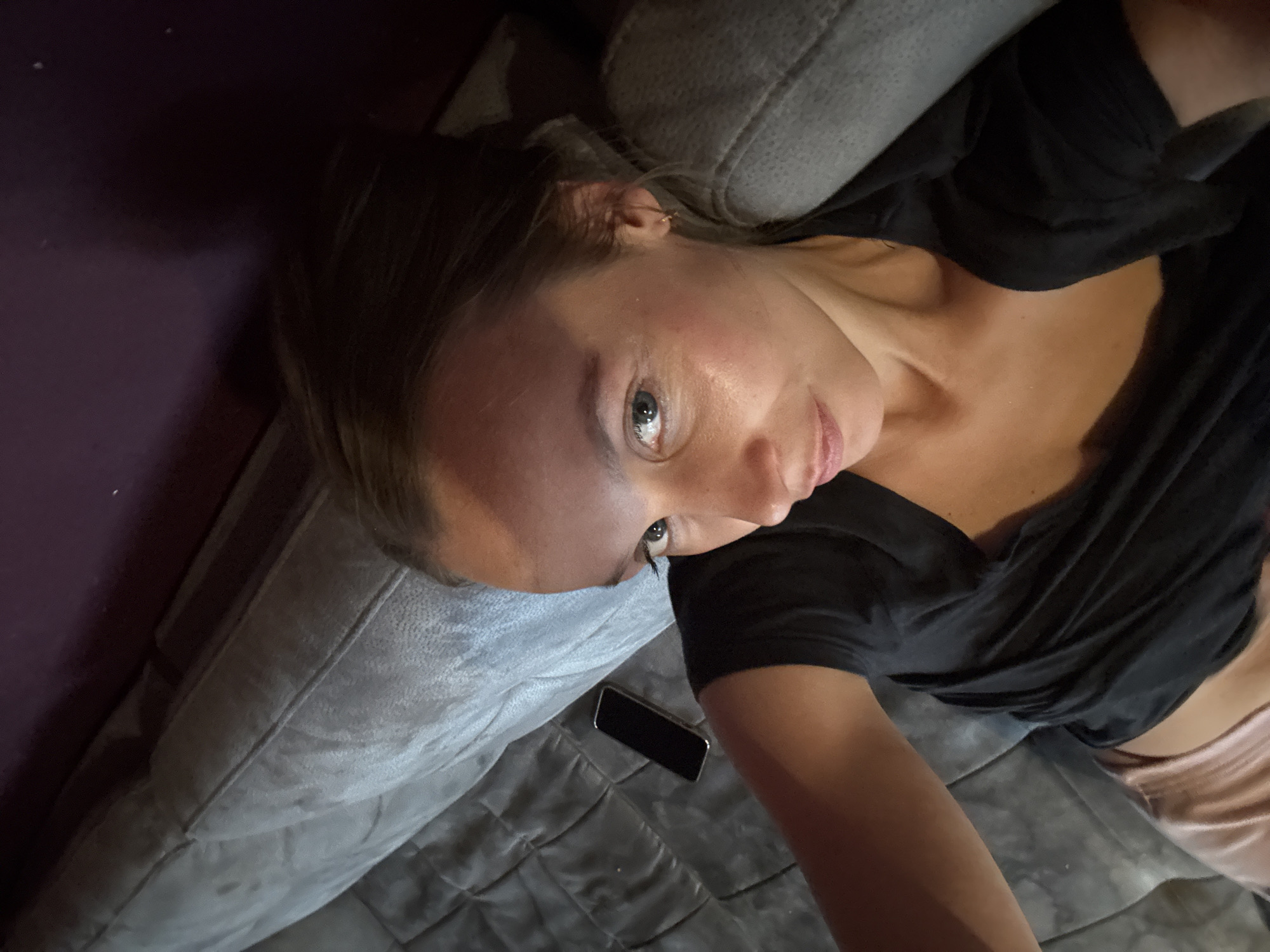 The image shows a woman lying on a grey couch in a dimly lit room. She is wearing a black t-shirt and pink shorts. Her hair is pulled back, and she is looking towards the camera. A smartphone is visible on the couch next to her. The background is a dark purple wall. The scene appears to be an indoor domestic setting, likely a living room or bedroom. There are no visible indicators that this location is in Überherrn, Germany.
