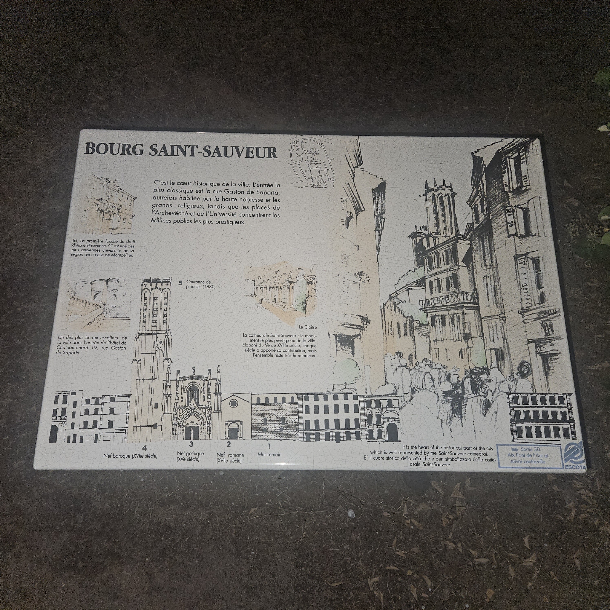 This is a photograph of a plaque depicting the historical center of Bourg Saint-Sauveur in Rousset, France. The plaque features a detailed black and white illustration of the city's architecture, including a prominent cathedral, other buildings, and a crowd of people. Text on the plaque describes the historical significance of the area, mentioning the rue Gaston de Saporta, the Archbishop's Palace, and the University. It also highlights specific architectural elements such as a Roman wall, Romanesque and Gothic naves, a Baroque nave, and the cathedral's pinnacles, dating from 1880. Smaller illustrations provide close-ups of the first law faculty of Aix-en-Provence and a staircase in the Hôtel de Chateaurenard. A section of text is also present in English and Italian, reiterating the importance of the Saint-Sauveur cathedral as the heart of the city. A blue logo with the text "Sortie 30. Aix Pont de l'Arc et suivre centre-ville ESCOTA" suggests an exit and directions within the city. The plaque appears to be made of ceramic tile, with a slightly aged appearance. The surrounding area is dark and appears to be outdoors, possibly in a park or garden, with some foliage visible at the top right.