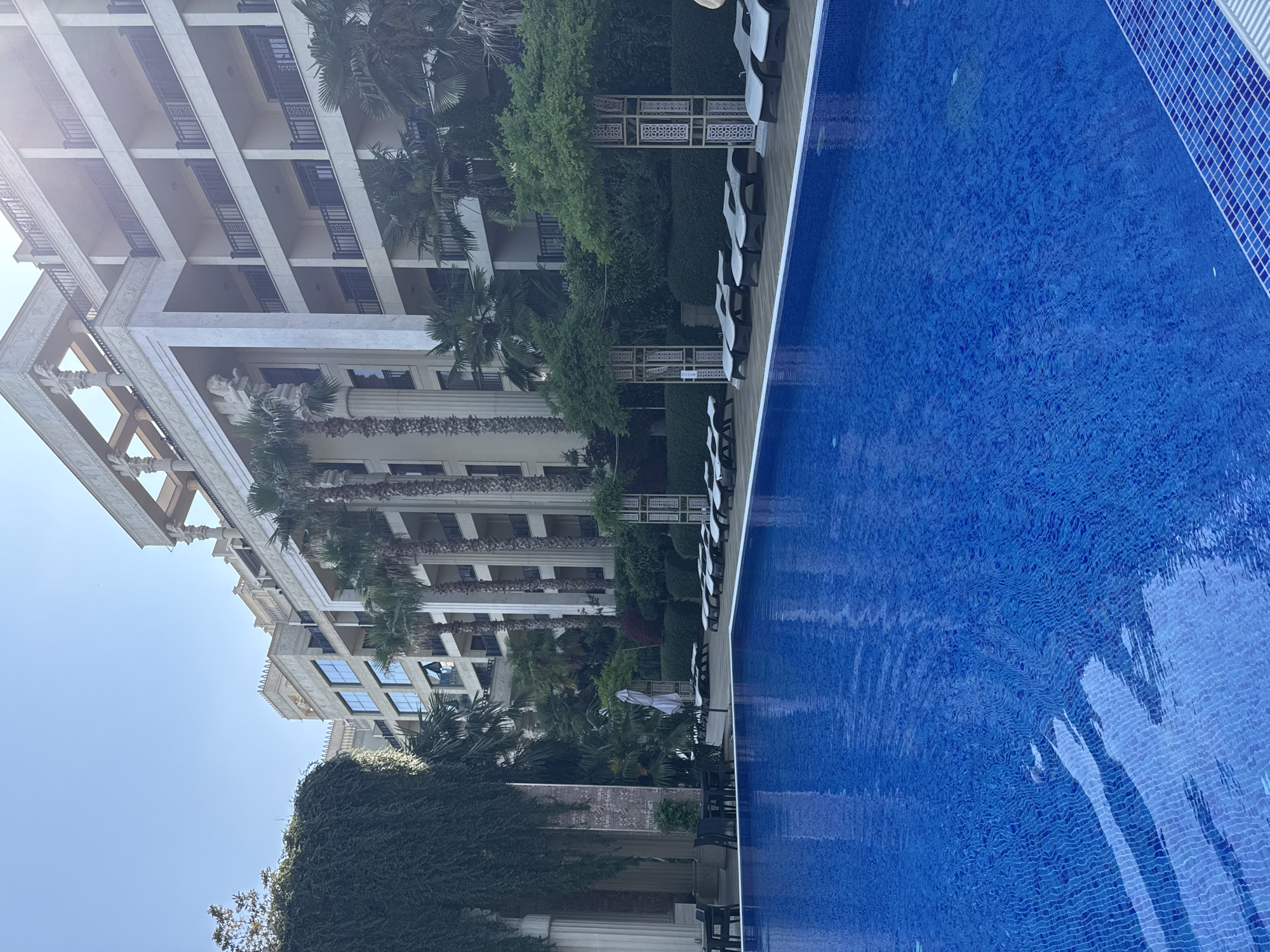 This image captures a bright, sunny outdoor swimming pool area, likely part of a hotel or resort. A large, multi-story building with a grand, ornate facade dominates the background. This building features numerous balconies with dark railings, classical columns, and decorative architectural details, suggesting a luxurious setting.
The foreground is occupied by a large, deep blue swimming pool with visible mosaic tiles at the bottom, and the water surface shows gentle ripples and reflections from the bright sunlight. Along the edge of the pool, a light-colored deck is lined with several lounge chairs and small tables, all arranged for relaxation.
Lush greenery, including numerous palm trees and manicured bushes, fills the space between the pool area and the building. Decorative white trellises with geometric patterns are interspersed within the landscaping. A single white umbrella is partially visible amidst the foliage.
The clear, bright sky, visible in the upper left, indicates a sunny day, likely morning or early afternoon. No people are present in the scene, conveying a sense of peaceful tranquility. The overall atmosphere suggests a high-end leisure or vacation destination in a warm climate. No discernible text is visible on any objects in the image.