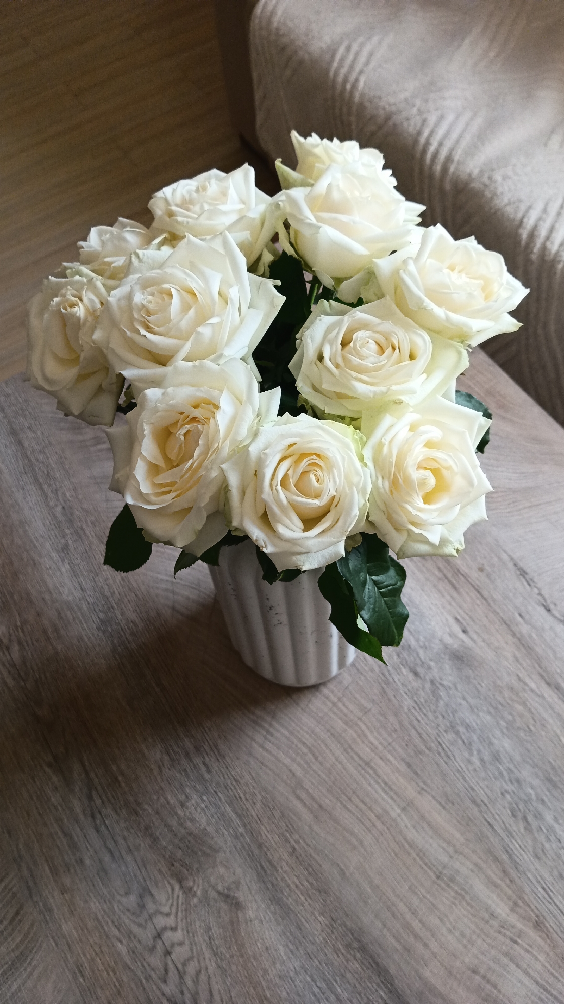 A vase of white roses sits on a wooden table, with a textured beige couch in the background. The roses are in full bloom and are arranged in a bouquet. The vase is white and ribbed. The table has a wood grain pattern. The couch appears to be made of fabric and has a plush texture. The lighting suggests an indoor setting, likely a home or a studio. There are no people or animals visible in the image. The scene is static and peaceful. The specific location in Ashtarak, Armenia, is not identifiable from the visual information provided in the image.