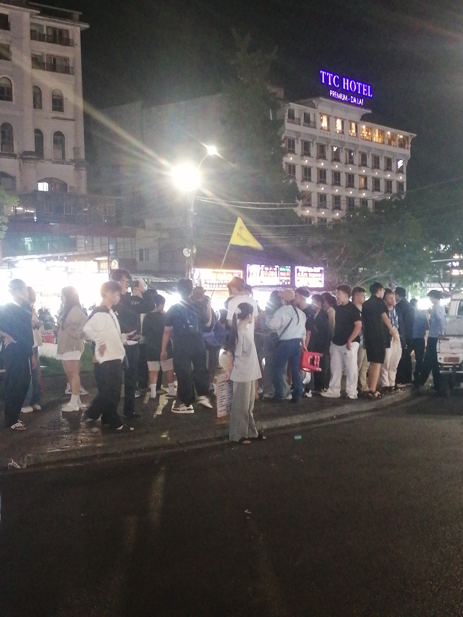 The image depicts a street scene at night in Da Lat, Vietnam. A group of approximately 20 people are standing on the sidewalk near a street. The street is wet. Buildings are visible in the background. A yellow flag is visible above the crowd. A sign for "TTC Hotel Premium Da Lat" is lit on a multi-story building. A white pick-up truck is parked on the street at the edge of the crowd. People are dressed in a variety of casual clothing. Some are talking. A streetlight is bright.