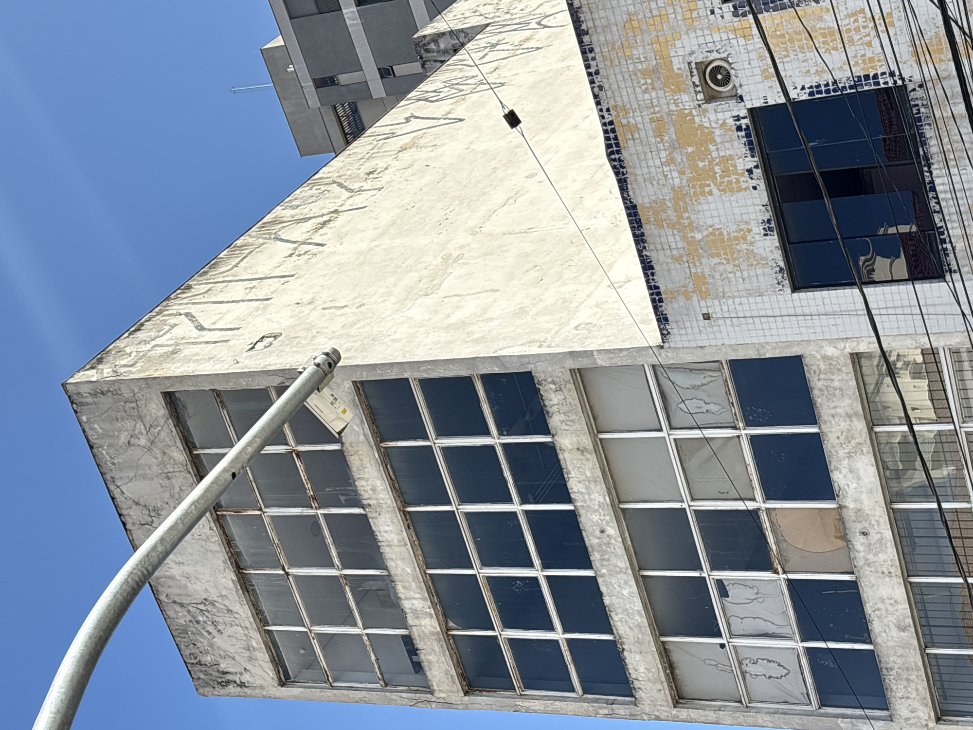 The image depicts an aged, six-story concrete building with a facade of large, multi-pane windows. The building appears to be in a state of disrepair, with visible cracks in the concrete and some windows covered with opaque material or appearing broken. The lower floors are adorned with a tiled pattern, partially weathered and discolored. Electrical wires are strung across the facade. In the foreground, a thick, metallic pipe arches diagonally across the frame, obscuring part of the building and sky. The background reveals a clear blue sky and a glimpse of another, more modern building. The scene is situated in the city of São Paulo, Brazil.