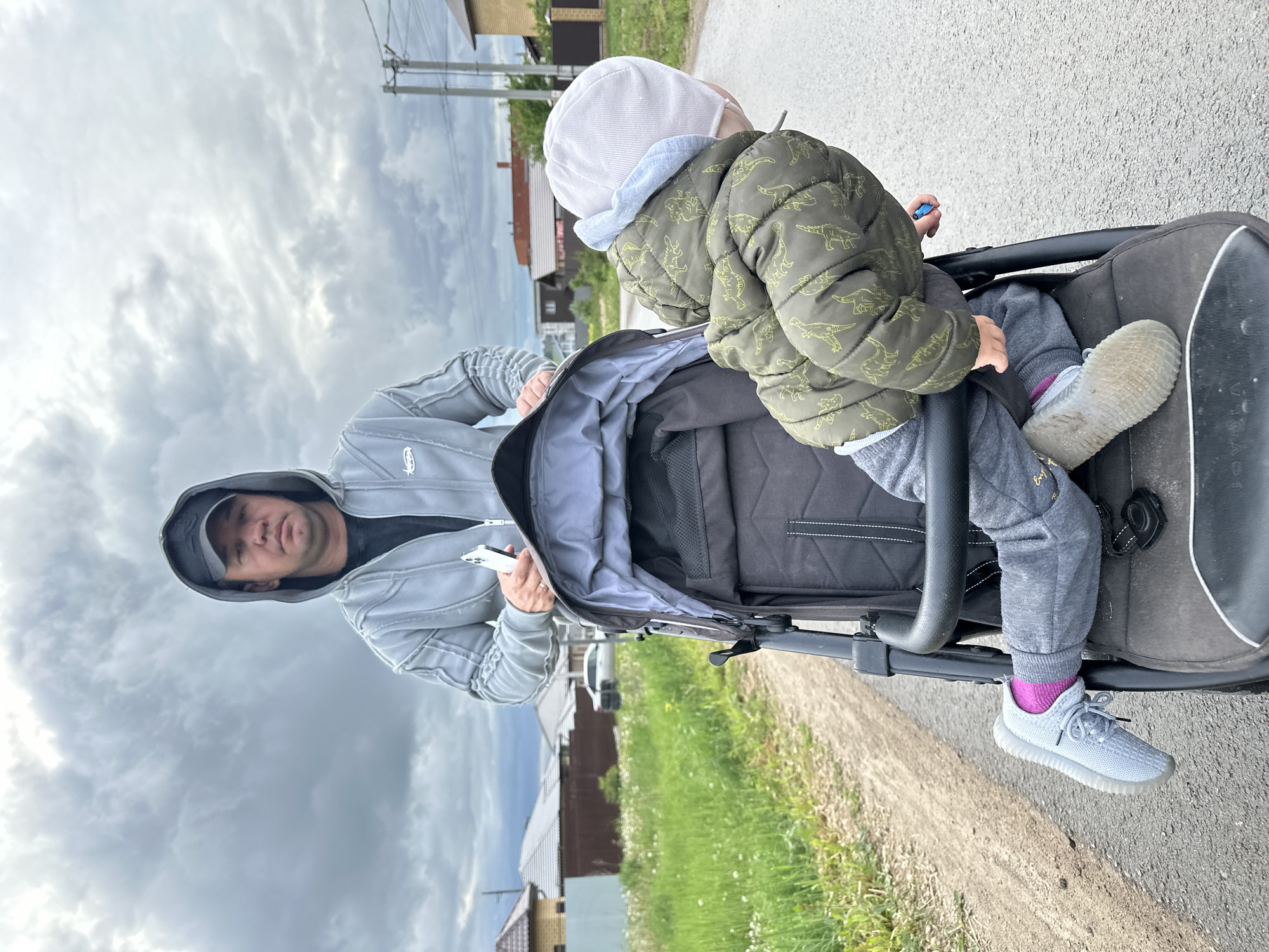 A man in a grey hooded jacket holds a smartphone, pushing a stroller with a child wearing a dinosaur-patterned jacket and a hat on a paved road. The location appears to be in Эстачи, Russia based on the visible architecture. The sky is overcast with grey clouds.