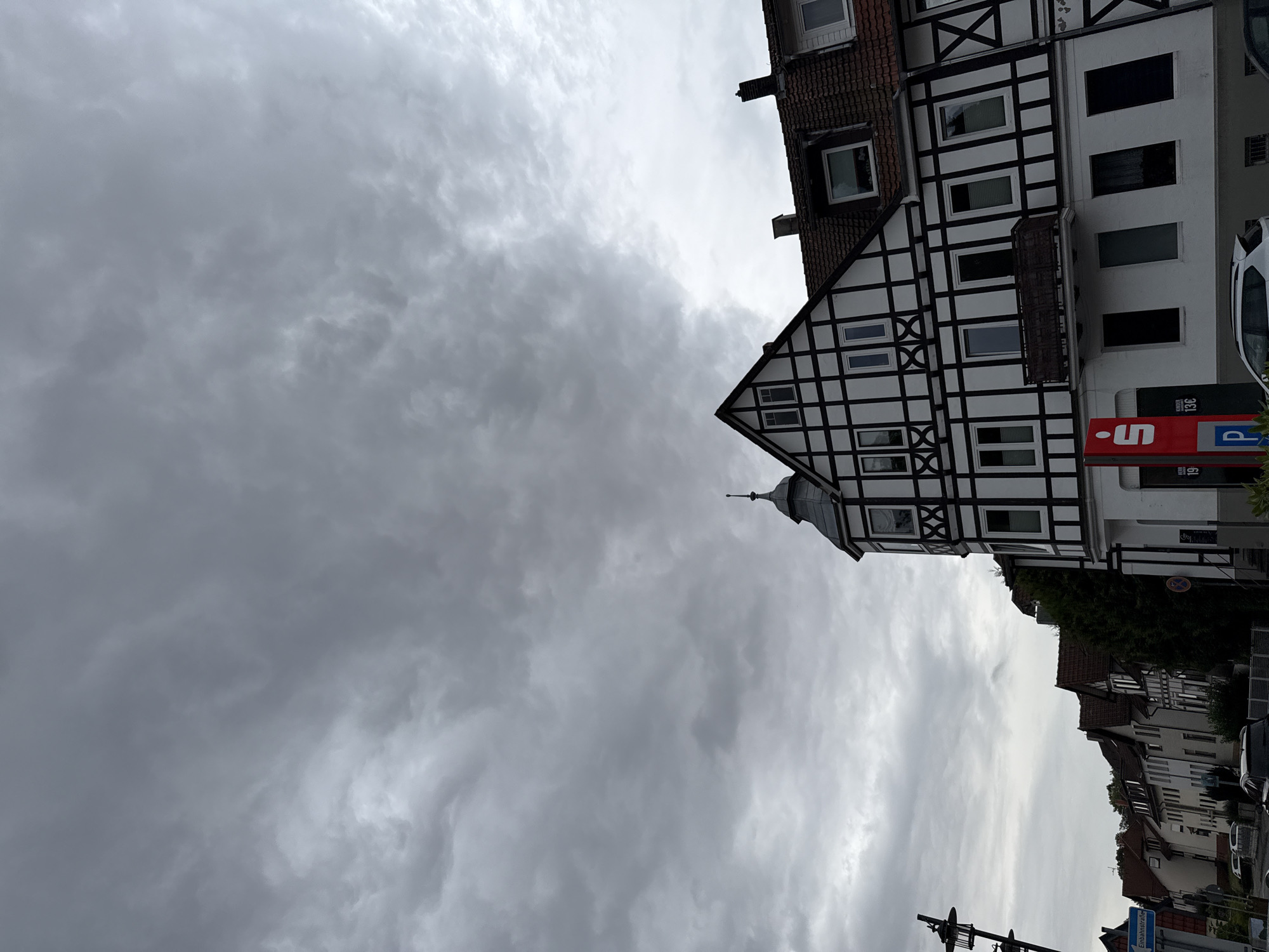 The image shows a street scene in Bad Sachsa, Germany, under a cloudy sky. On the right is a multi-story building with a white facade and black half-timbering, characteristic of German architecture. It features multiple windows, some with shutters, and a small balcony. A red sign with a white "S" logo, likely for Sparkasse (a German savings bank), is visible on the ground floor. To the left of this building, a row of residential houses stretches into the distance, also featuring pitched roofs. A blue directional sign on a pole indicates "Einbahnstraße" (one-way street). A black lamppost stands on the left side of the image. Cars are parked along the street and on the right side of the building. The ground is paved, suggesting a sidewalk and road. The overall atmosphere is overcast and somber.