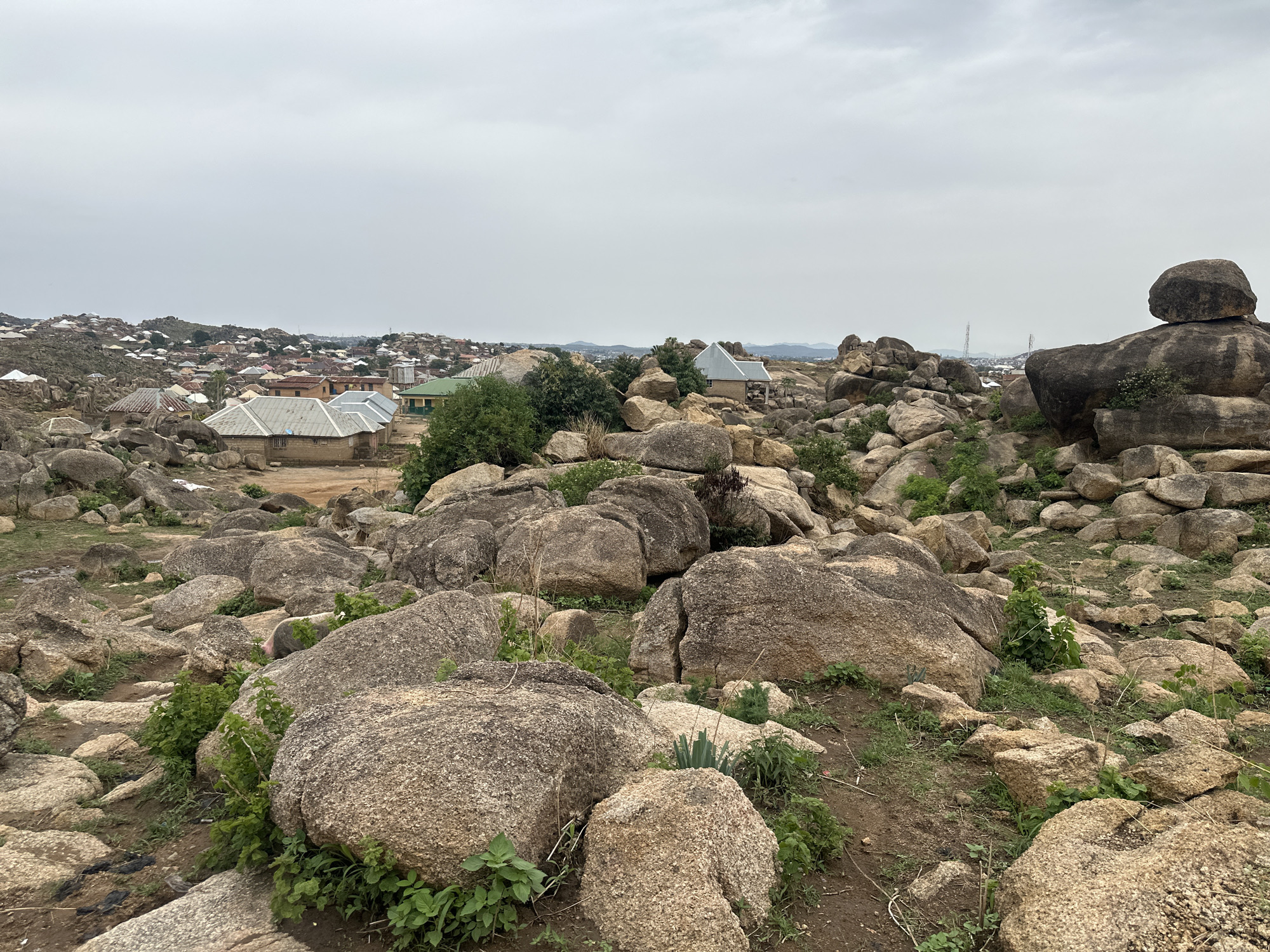 The image depicts a rocky landscape in Jos, Nigeria. Large, rounded granite boulders dominate the foreground, interspersed with sparse vegetation consisting of small shrubs and grass. In the middle distance, a collection of buildings, some with corrugated iron roofs, indicates a residential area. The structures vary in size and color, suggesting a mix of dwelling types. In the background, a hazy sky covers the city. Power lines are visible in the background.