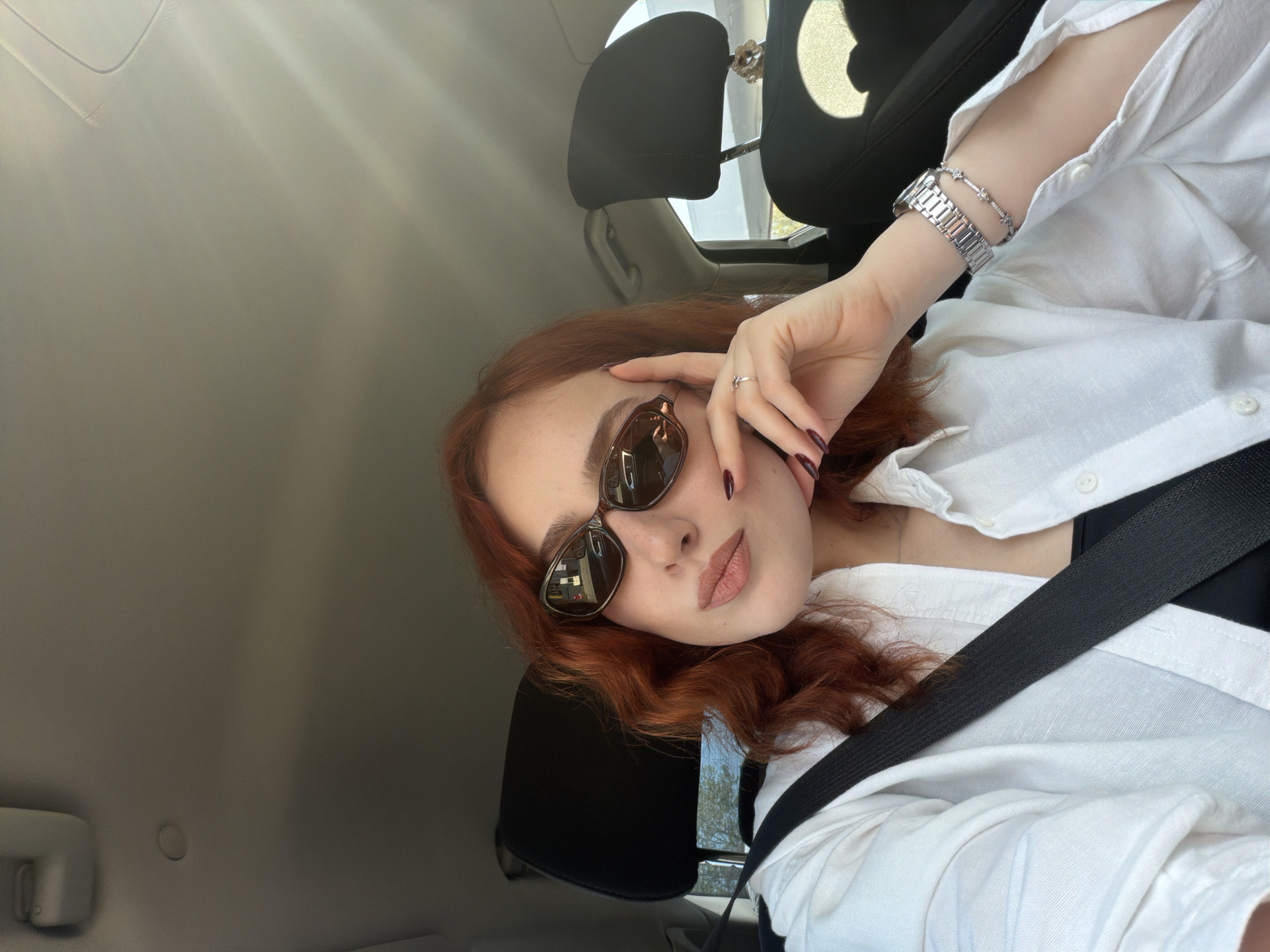 The image displays a woman with reddish-brown, wavy hair, captured in what appears to be a selfie taken inside a car in Tbilisi, Georgia. She is seen from a slightly overhead, tilted perspective, wearing dark sunglasses that partially obscure her eyes. Her lips are painted in a soft, nude-pink shade.
She is dressed in a white, collared button-up shirt, possibly over a black top, and is wearing a black seatbelt across her chest. Her left hand, adorned with dark red nail polish and a subtle ring on her index finger, is positioned near her face, gently touching the frame of her sunglasses. On her left wrist, she wears a silver-toned watch with a metal strap and a delicate sparkling bracelet.
The setting is the interior of a car, with light-colored headrests and a matching ceiling visible in the background. Rays of sunlight stream into the vehicle from the top left, indicating it is daytime and sunny outside, casting subtle patterns of light and shadow within the car. Reflections in her sunglasses offer a blurred glimpse of outdoor elements like trees and a building. No visible text is present in the image. The overall mood is relaxed and casual.