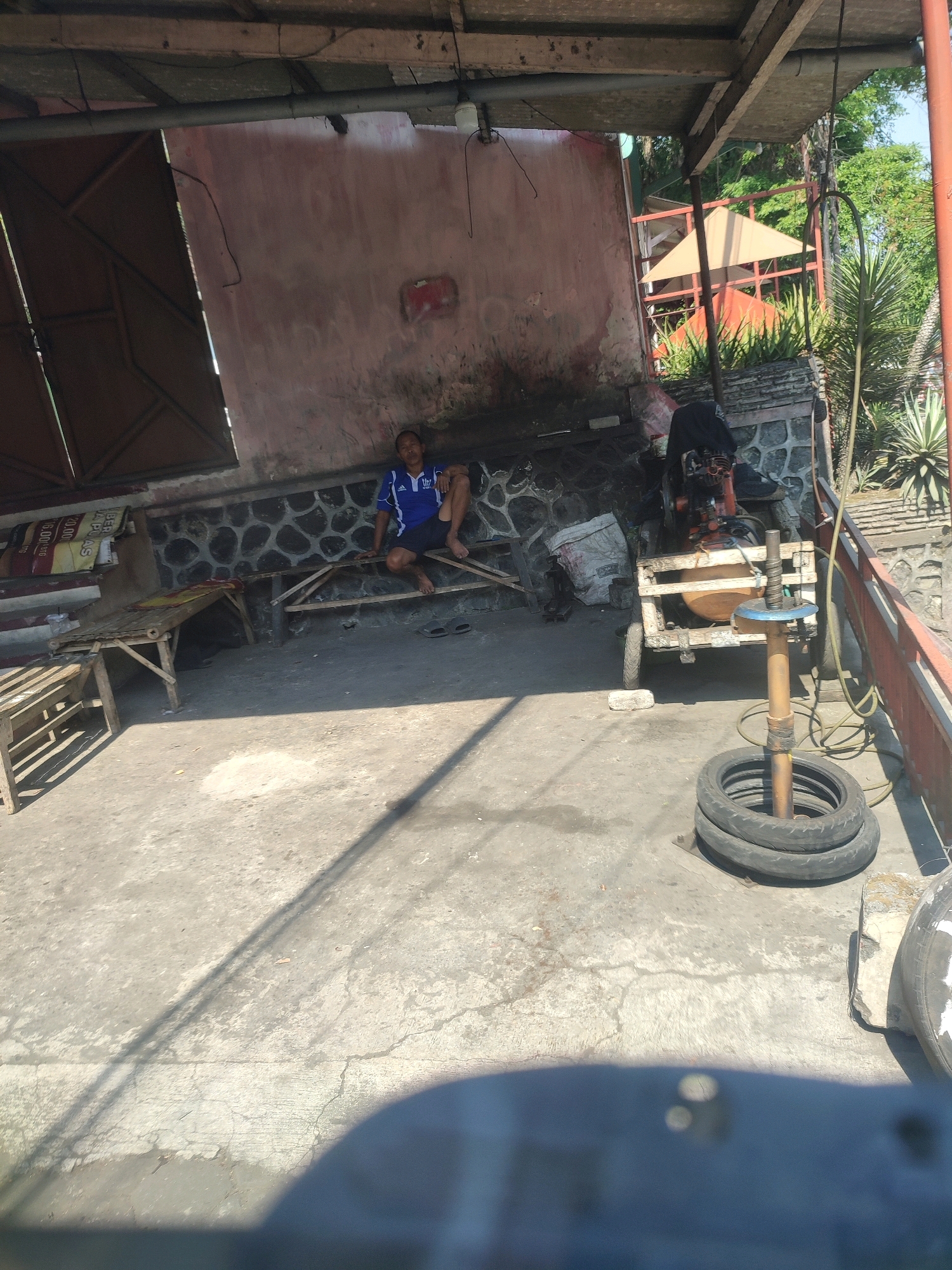 In Tonoboyo, Indonesia, a man is sitting on a wooden bench under a shaded, open-air structure. The structure has a corrugated metal roof and a stone wall. The man is wearing a blue t-shirt and dark shorts. In the foreground, there is a concrete floor with visible cracks. To the right, a wooden cart with a motor is positioned, and two tires are stacked on the ground next to it. Behind the cart, there are plants and trees, suggesting an outdoor setting. A large, pinkish wall serves as the backdrop for the scene.