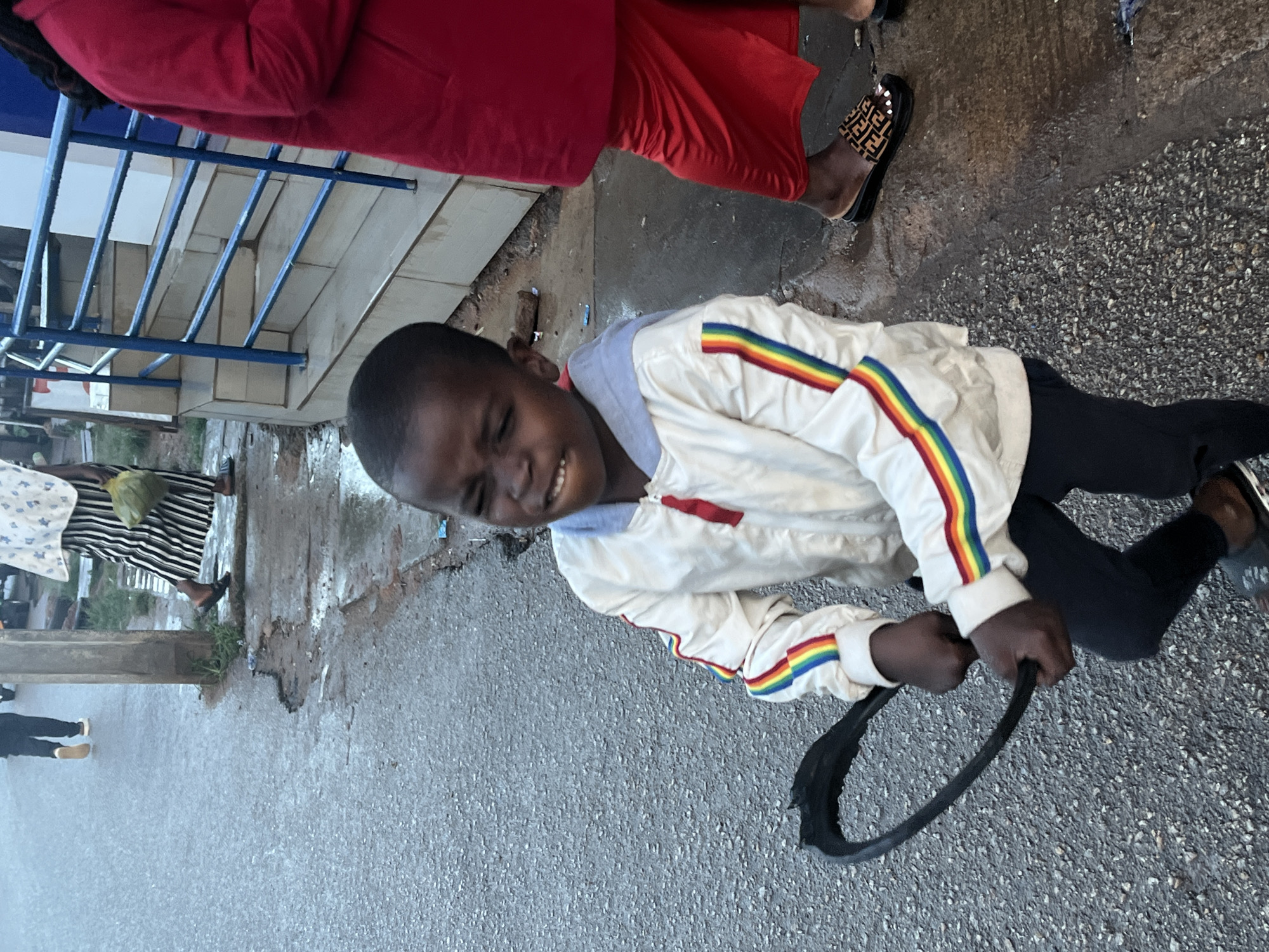 A young Black boy, wearing a white jacket with rainbow stripes on the sleeves and black pants, stands on a wet asphalt street in Jos, Nigeria. He is holding a black, circular object, possibly a tire inner tube, in both hands and appears to be straining or making a distressed facial expression. To his right, a woman in a bright red dress and black slide sandals with a brown and black pattern stands with her legs visible from the knees down. Behind the boy, other individuals are present, including a person in a white and patterned garment and striped skirt, and another person walking away on the left side of the frame, wearing dark pants and light-colored shoes. The environment appears to be an urban setting with a sidewalk, a metal railing, and a building in the background. The ground is wet, suggesting recent rain.