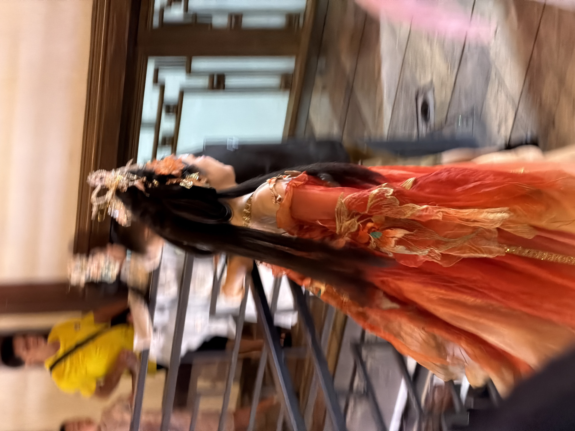 In Yuzhong District, China, a blurred image captures a person in traditional Chinese attire, possibly a Hanfu, with a vibrant orange and red layered skirt and ornate gold headwear. The person is seen from the side, with long dark hair cascading down their back. They are standing on a staircase with metal railings. In the background, another individual in a yellow t-shirt is visible, and further back, a wooden lattice-like structure suggests an interior or architectural setting. The motion blur indicates movement within the scene, possibly during a performance or parade.