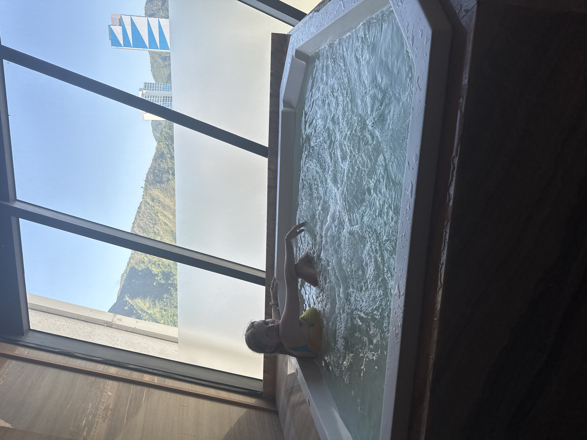 A young woman with blonde hair is sitting in a large, white, bubbling hot tub. She is wearing a yellow and blue bikini. Her arms are resting on the edge of the tub and she is looking at the camera. The hot tub is located indoors, with a large window behind it offering a view of a city skyline and green mountains under a clear blue sky. The window is framed by dark wood paneling and angled dark beams. The hot tub itself is filled with clear, turbulent water, and the edges are wet with droplets. The overall setting appears to be a hotel or resort room.