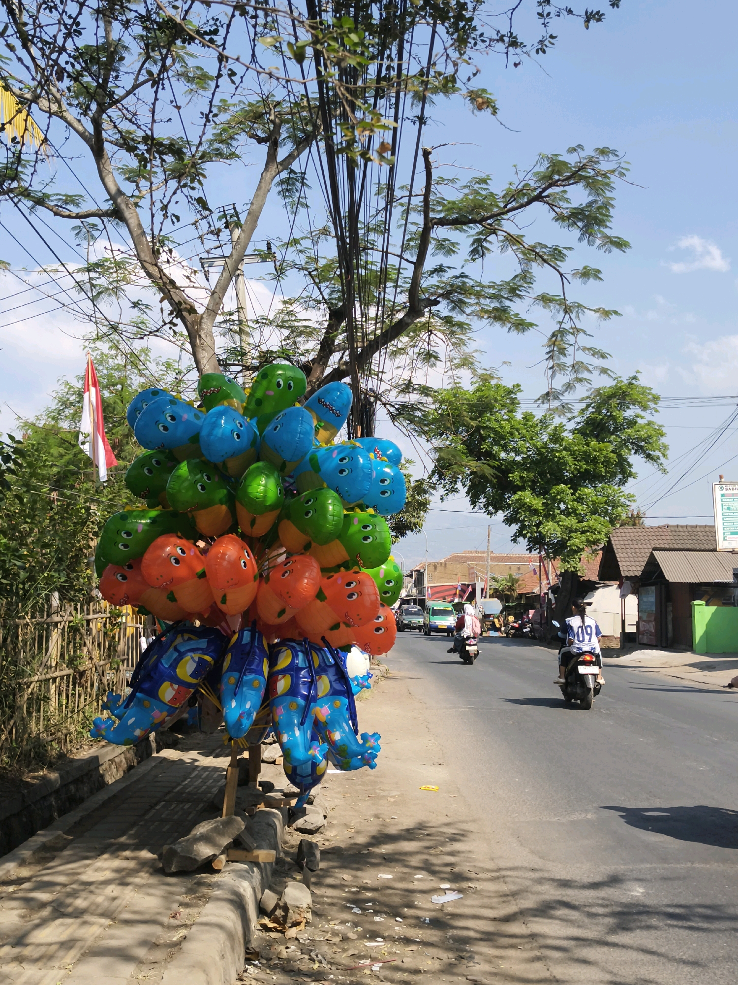 A vendor is selling balloons along a street in Majasetra, Indonesia. A colorful assortment of dinosaur-shaped balloons, primarily blue, green, and orange, are tied to a wooden stand on the sidewalk. The balloons are arranged in a cluster, with some shaped like individual dinosaurs and others like pairs of dinosaurs. The street is paved and appears to be a two-lane road with traffic, including motorcycles and a small van. The buildings on the side of the street are typical of a rural or semi-urban Indonesian setting, with simple structures and tiled roofs. A red and white Indonesian flag is visible in the background, adding to the sense of place. The sky is clear with some scattered clouds, and the sunlight casts shadows on the ground. The scene is bustling with activity, typical of a market street.