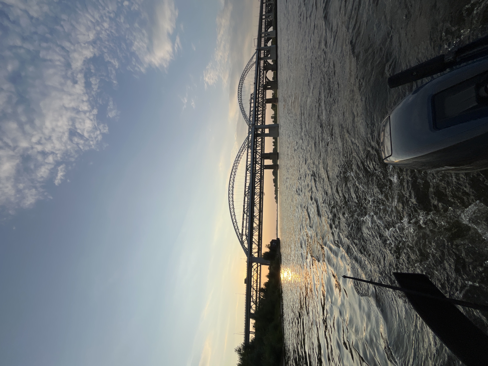 A wide shot captures a vast river in Nizhny Novgorod, Russia, at sunset. The sky is a gradient of soft blues and oranges, with wispy clouds scattered across the horizon. In the distance, a grand arched bridge stretches across the water, its structure silhouetted against the fading light. The sun's reflection shimmers on the rippling surface of the river. In the foreground, the stern of a boat is visible, with an outboard motor and a paddle. The water behind the boat is disturbed, showing the wake of its movement. The overall scene depicts a serene evening on the river, with the iconic bridge serving as a prominent landmark.