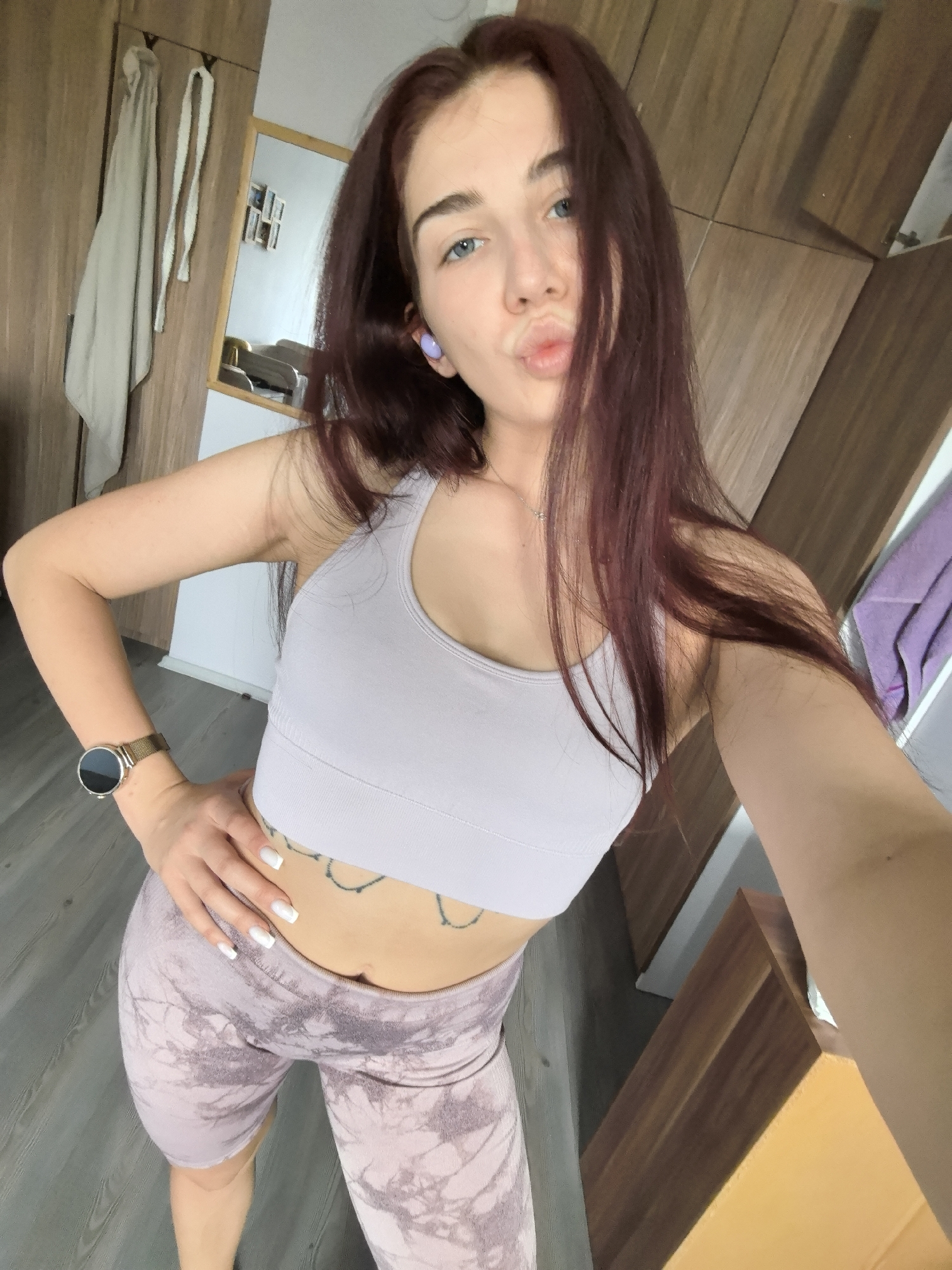A young woman takes a selfie in an indoor setting, likely a residence in Stara Zagora, Bulgaria. She has long, dark reddish-brown hair, blue eyes, and is making a playful "duck face" expression towards the camera.
She is wearing a light lavender-colored ribbed sports bra and matching tie-dye patterned biker shorts. A black line-art tattoo is visible on her lower abdomen. On her left wrist, she sports a smartwatch with a gold mesh band, and a light purple wireless earbud is visible in her left ear. She also wears a delicate silver necklace and has white, square-tipped fingernails. Her right hand is visible, holding the camera, while her left hand is placed on her hip.
The background features warm-toned wooden wardrobes or cabinets. On the left side of the frame, two light-colored garments, possibly robes or scarves, hang on hooks. A rectangular mirror with a wooden frame is visible behind her, reflecting part of a room that appears to contain furniture and framed pictures. On the far right, a hint of purple fabric or a towel is visible. The flooring is light gray, resembling wood laminate or planks. The scene is brightly lit, indicating it is daytime. The woman projects a confident and playful demeanor.