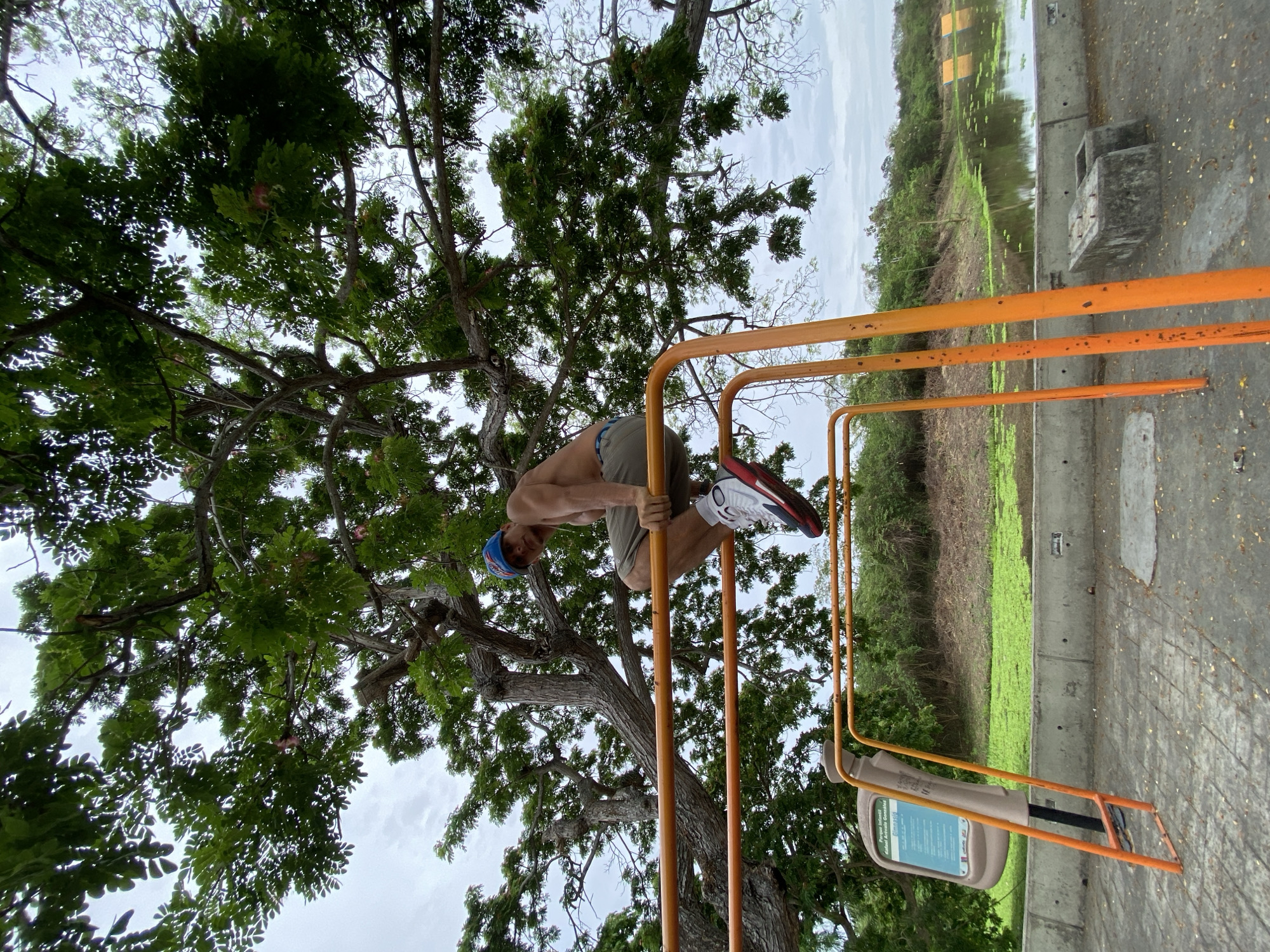A man in athletic wear is executing a gymnastic maneuver on parallel bars in Río Chico, Venezuela. He wears a blue cap, khaki shorts, a white sock, and a red and white athletic shoe. He hangs upside down while holding onto the parallel bars. The bars are bright orange and set in a park-like setting. Behind the bars and the man, a large tree with lush green foliage dominates the left side of the frame. Behind the bars on the right side, there is a body of water covered with vegetation, suggesting a wetland or marsh. The weather appears overcast.
