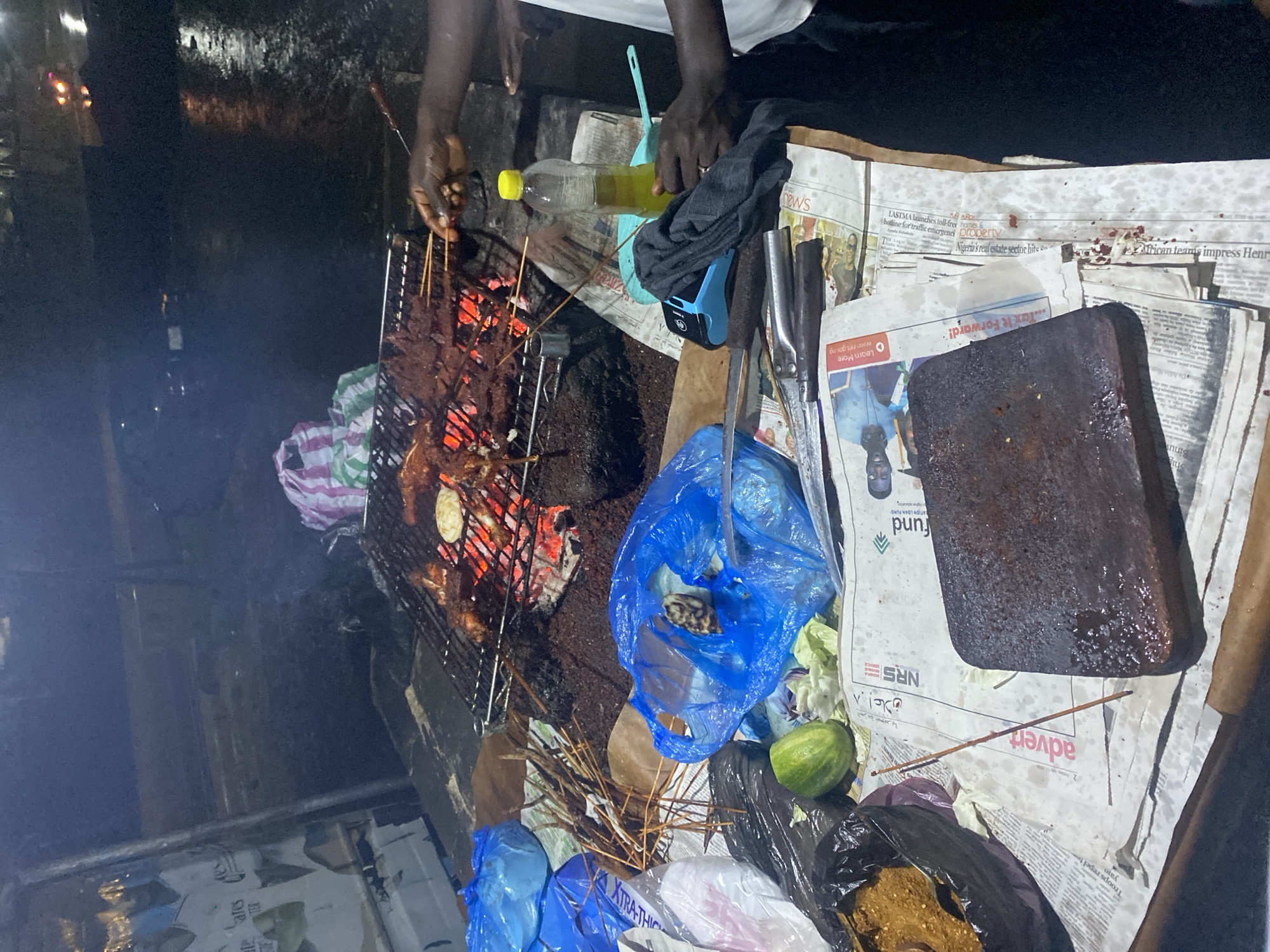 This is an outdoor nighttime scene in the city of Karum, Nigeria, depicting a food stall. The stall is characterized by a grill laden with various meats, some of which are skewered and being cooked over glowing embers. A person's hands are visible tending to the grill, suggesting they are the vendor. In the foreground, a collection of items is spread out on a surface covered with newspapers. These items include plastic bags, a cucumber, a bottle of oil, a cutting board, and various utensils. The overall impression is that of a busy, informal food preparation area, likely a common sight in urban Nigerian settings at night.