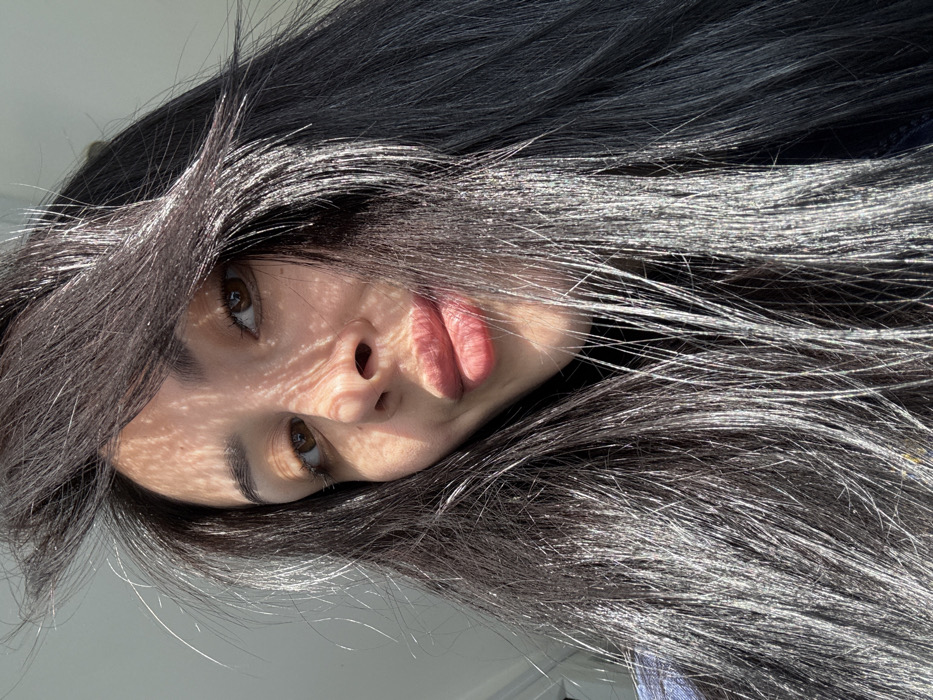 A close-up, upward-angled portrait shot depicts a female subject with long, dark hair, dark-brown eyes, and full, pink-tinted lips forming a slight pout. She is looking directly at the camera. Strong, directional sunlight from the upper left casts distinct, patterned shadows, resembling a lattice or blind, across the left side of her forehead, cheek, and nose. The sunlight also creates prominent specular highlights on her dark hair, making sections appear shiny and silvered. A dark collar is visible at her neck, and a portion of a blue garment is seen on her left shoulder. The background is a brightly lit, out-of-focus, light-colored interior wall. The scene is located in the city of Molodezhnyi, Russia.