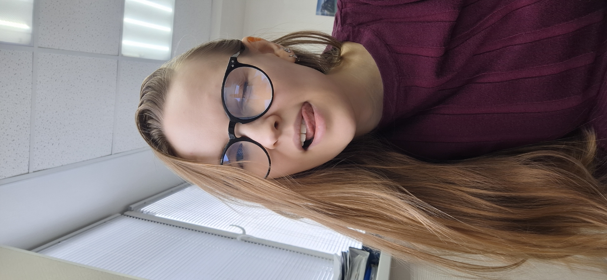 A female subject is depicted in a close-up, low-angle indoor portrait. She has long, light brown hair, is wearing black-rimmed circular glasses, and a dark burgundy ribbed long-sleeved top. Her left eye (viewer's right) is closed in a wink, and her tongue is partially extended from her open mouth. Multiple small earrings are visible on her right earlobe (viewer's left). The background features a white drop ceiling with visible rectangular fluorescent light panels in the upper right quadrant. To the upper left, a window fitted with horizontal white blinds and a white handle is partially visible. A stack of papers rests on a surface beneath the window. A framed picture, possibly a landscape, is discernible on the wall in the far right background. The specific location within the city of Chaikovsky, Russia, cannot be determined from the visual information provided, beyond it being an interior commercial or office environment.