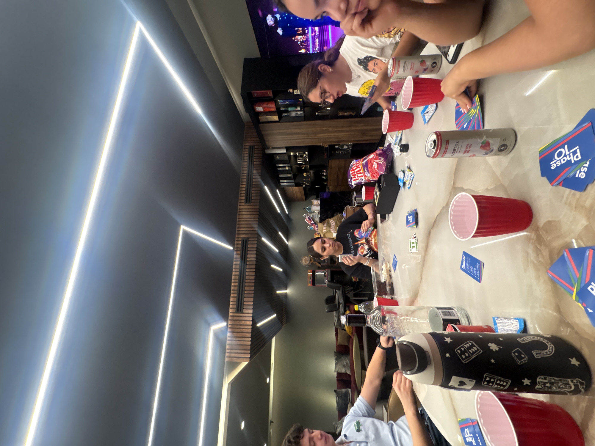 **Scene:** An indoor gathering in a modern, well-lit space.
**People:** Multiple individuals are seated around a table. They are engaged in a card game.
**Objects:**
* Playing cards (Phase 10)
* Red plastic cups
* Cans of Seagram's Escapes
* Snack bags
* Water bottles
* Television
* Shelf with miscellaneous objects.
**Actions and Interactions:** The individuals are playing cards, presumably in a social setting.
**Environment:** The space is characterized by contemporary design elements, including recessed lighting and wooden wall accents.
**Location:** The image was not taken in San Bernardino Tlaxcalancingo, Mexico.