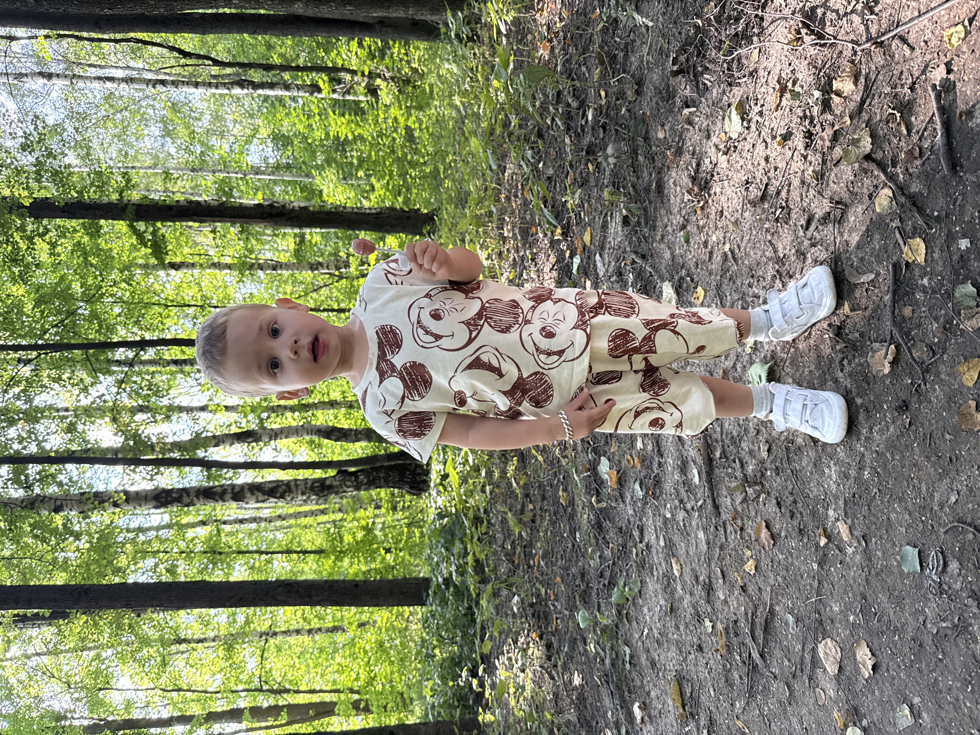 A young boy stands in a forest in Rusanovo, Russia, holding a lollipop. He is wearing a beige outfit with a brown Mickey Mouse pattern, white socks, and white sneakers. The boy has light brown hair and is looking at the camera with an open mouth. The forest floor is covered in dirt, fallen leaves, and twigs. Sunlight filters through the dense green foliage of the trees, creating a dappled light effect on the ground.