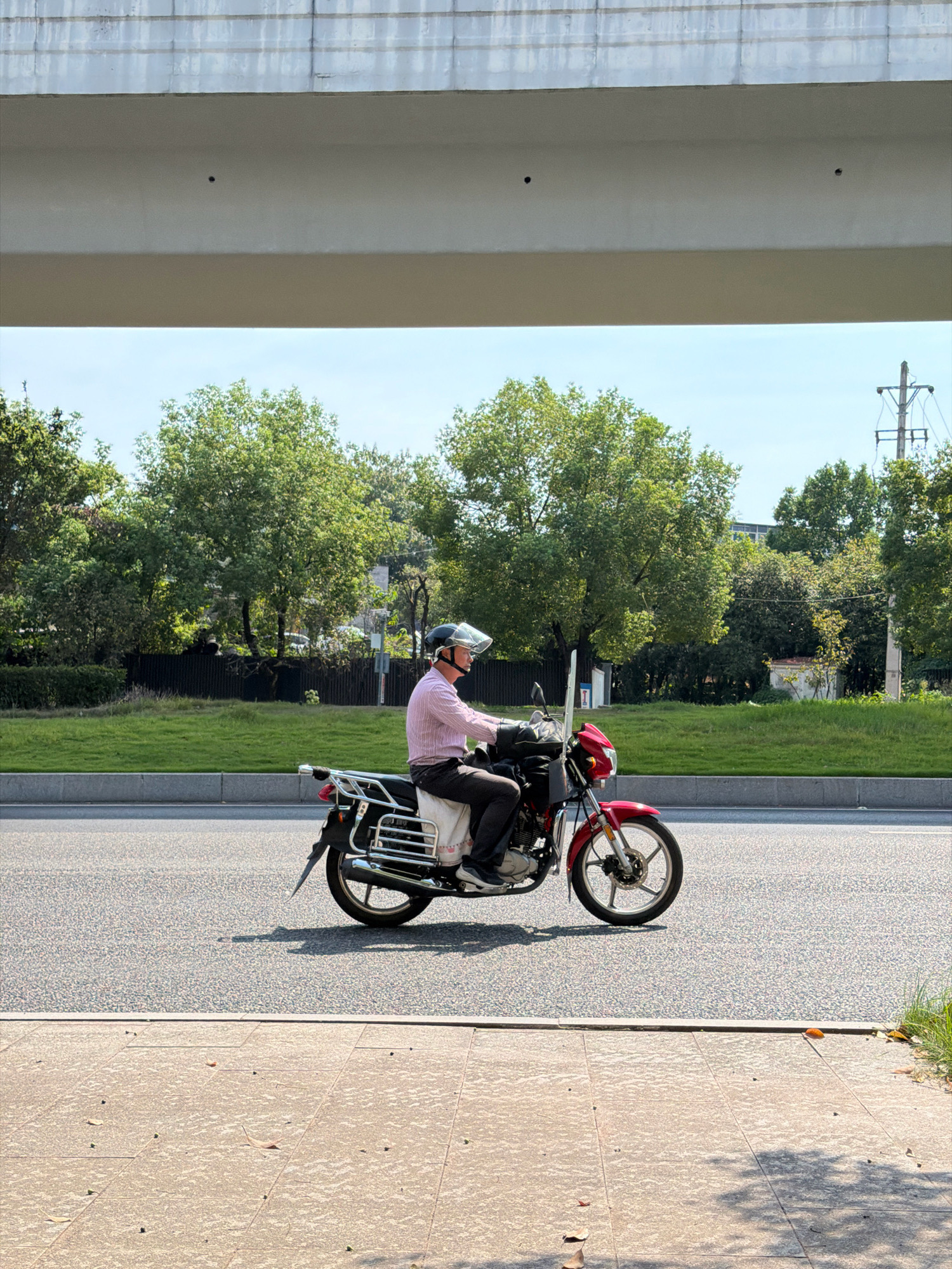 A man wearing a pink collared shirt, dark pants, and a helmet is riding a red and black motorcycle on an asphalt road. He is looking forward and appears to be in motion. The motorcycle has a chrome luggage rack on the back and a small windshield. The scene is outdoors, with lush green trees and grass in the background. A concrete overpass is visible in the upper portion of the image. The sky is clear and blue. The image was captured in the city of Jiangjin, China.