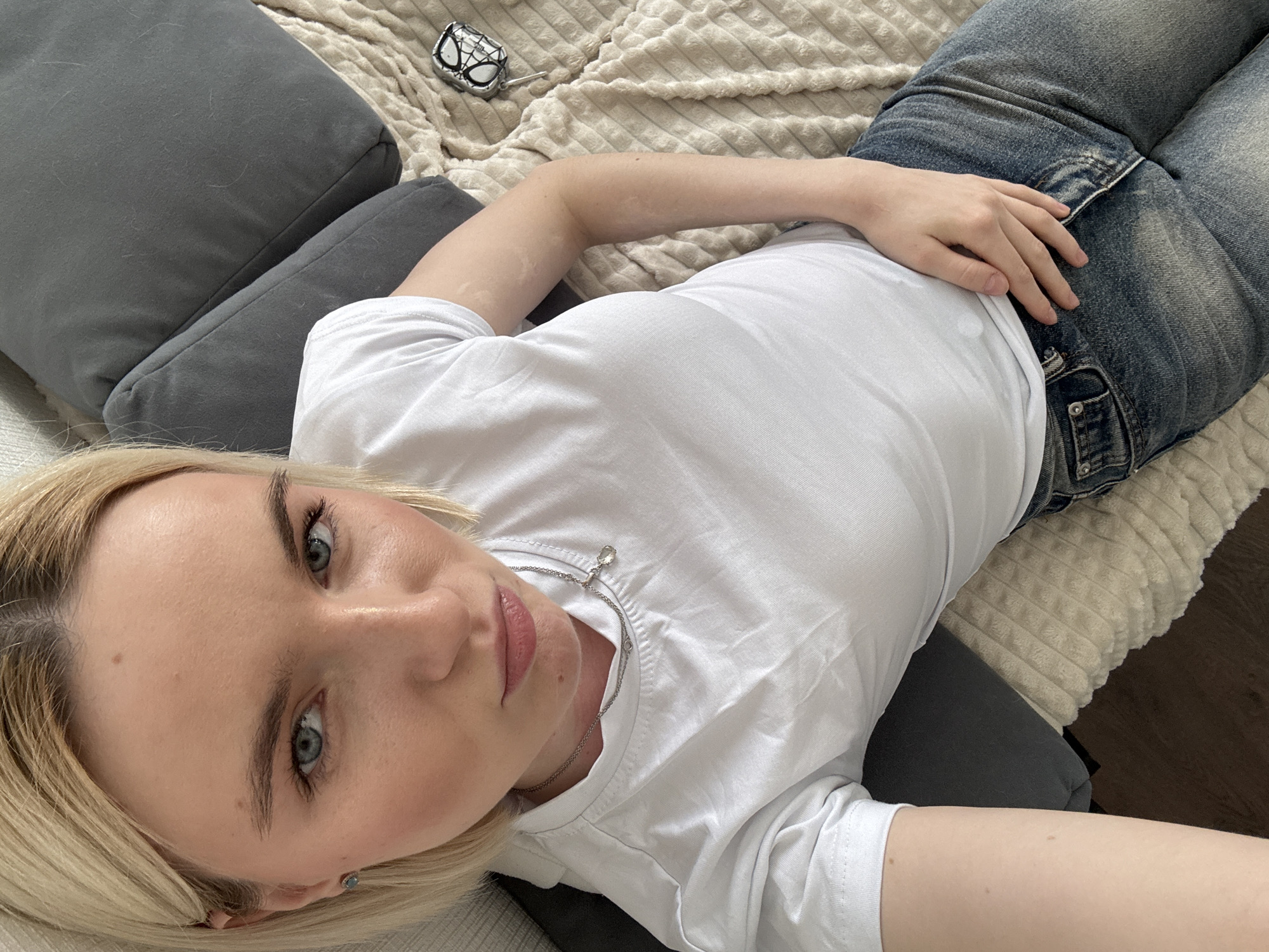 A young woman is lying on a bed or sofa. She is wearing a white t-shirt and blue jeans. Her blonde hair is visible on the left. The bedding is textured beige, and there are grey pillows. A small object resembling a spider-man mask is on the bed. The photograph appears to be taken from a high-angle perspective. The location cannot be determined as Vsevolozhsk, Russia.