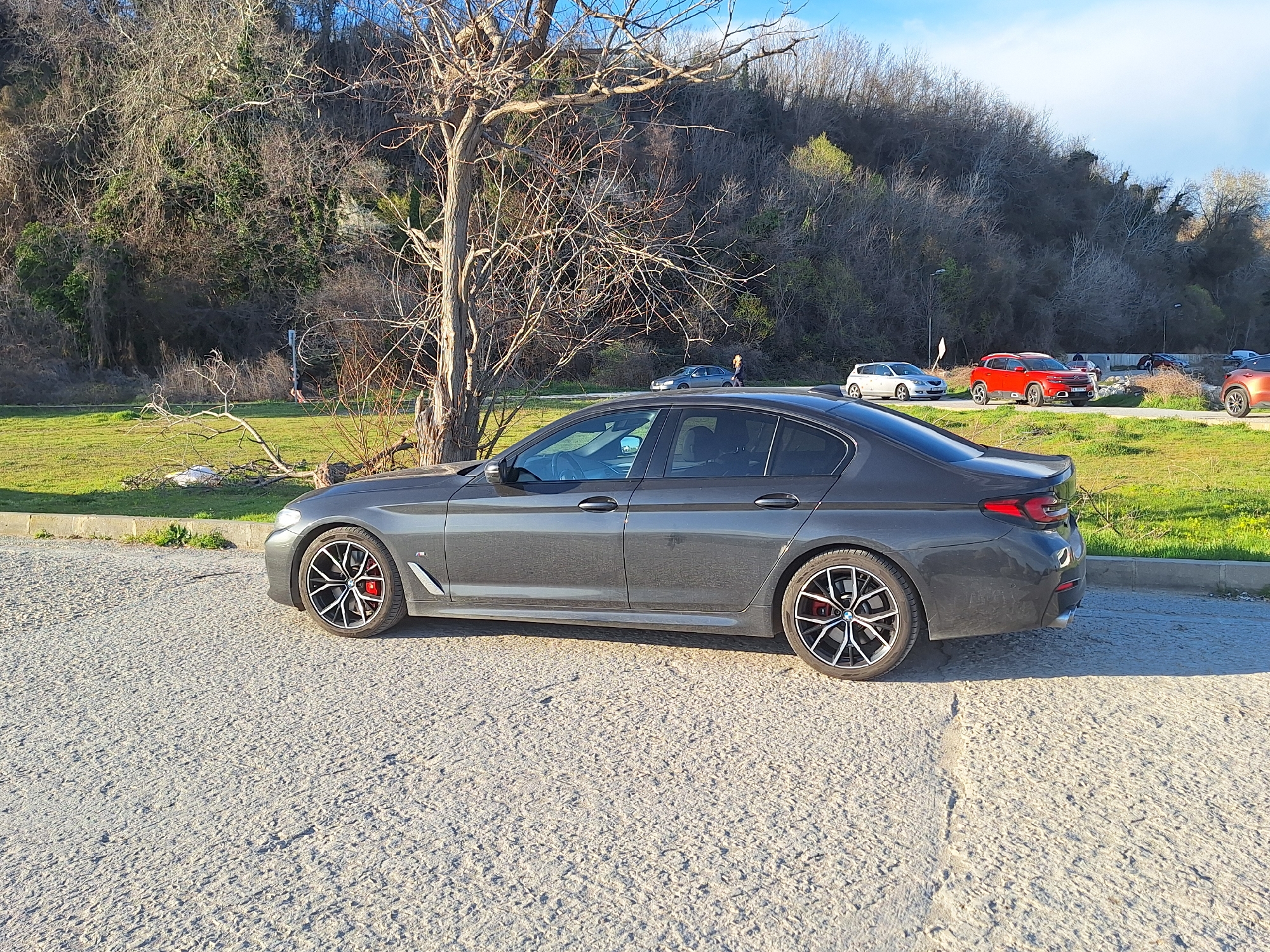 The image depicts a dark gray BMW 5 Series sedan parked on a rough, light-colored paved surface. The car has distinctive black and silver wheels with red brake calipers. It is parked in front of a grassy area with a leafless tree in the foreground and a densely wooded hillside in the background. In the distance, several other cars are visible, including a red SUV and a silver hatchback.
Given the distinctive landscape, it is highly probable that the location is the city of Varna, Bulgaria. The car is parked in a designated location. The weather appears to be sunny with a few clouds in the sky. There are no discernible people immediately visible in the foreground near the parked car.