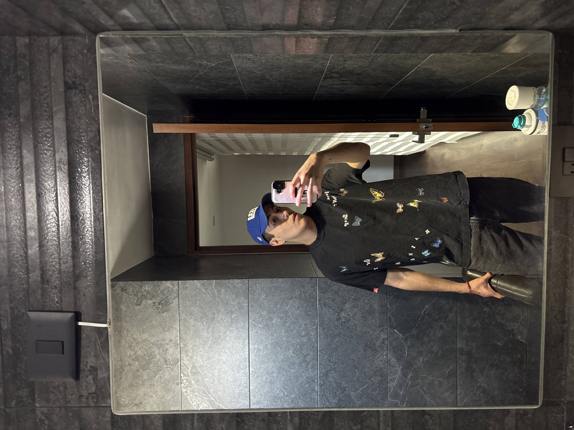 A mirror reflects a man in a bathroom-like setting. He is wearing a blue baseball cap, a dark t-shirt with butterfly designs, and dark pants. He is holding a phone in one hand and a dark-colored object in the other. The mirror is surrounded by dark grey tiles. The reflection shows part of the bathroom interior, including what appears to be a shower curtain. The presence of a light switch and toiletries further suggests a bathroom setting. The location is not discernible from the image.