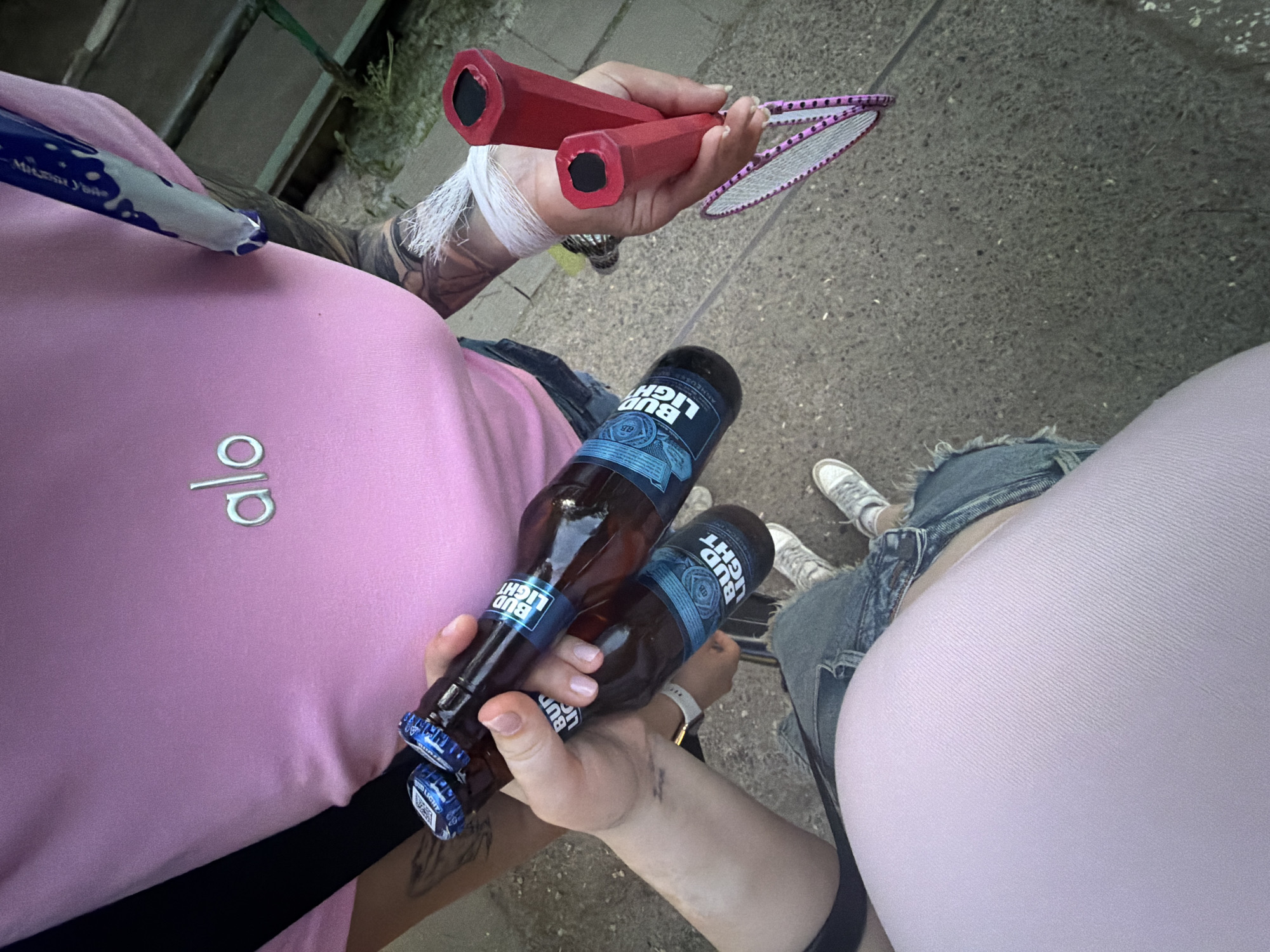 Two individuals are visible from an overhead perspective. The person on the left is wearing a pink shirt with the "alo" logo, and black pants. They hold two bottles of Bud Light beer in their left hand and a pair of red badminton racquets in their right hand. The other person is wearing a pink shirt with jeans and grey sneakers. The setting appears to be an outdoor location. It is not possible to determine the location within the city of Chaikovsky, Russia, from this image alone.