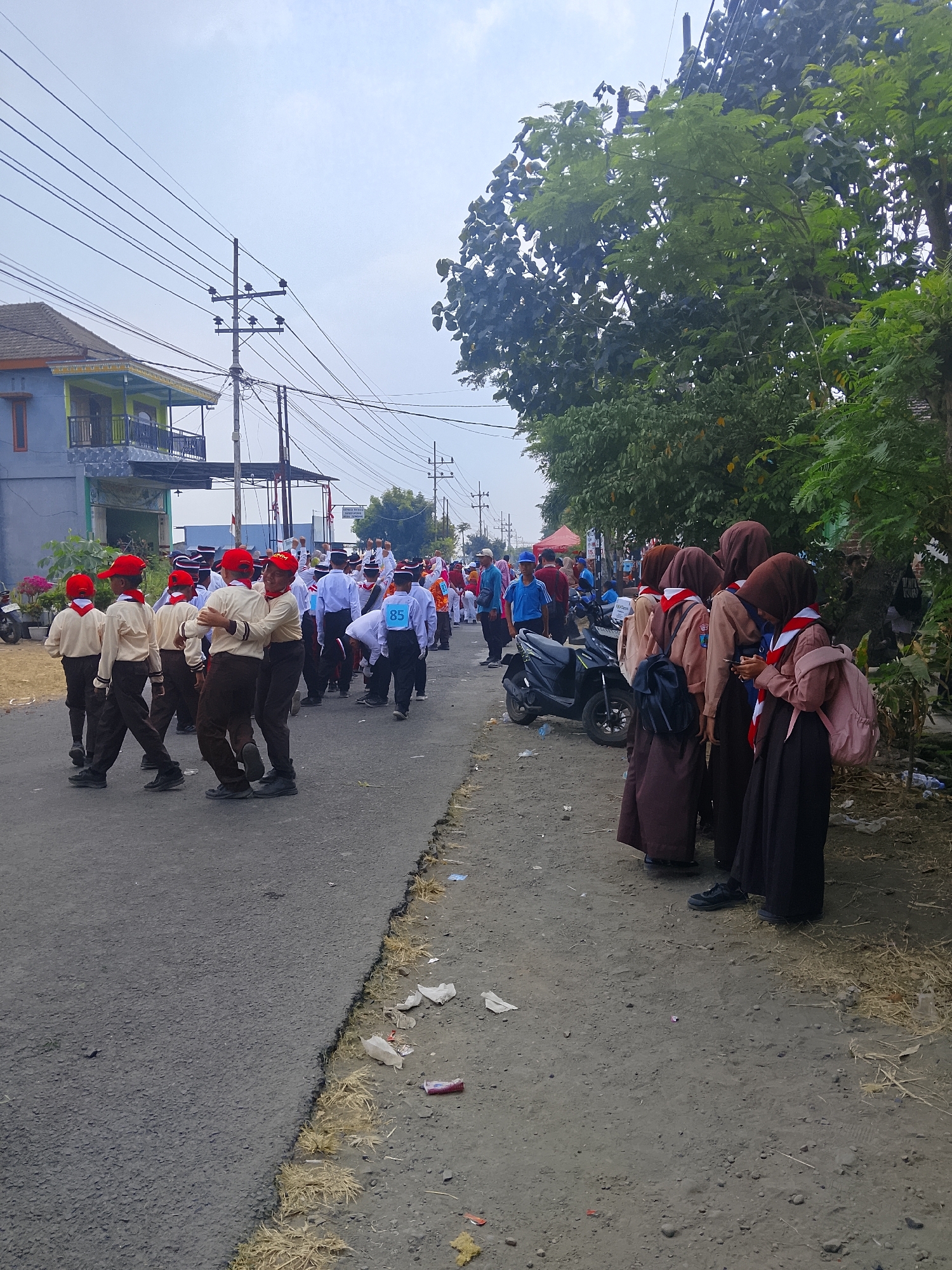 In Juwono, Indonesia, a group of uniformed schoolchildren, likely participating in a parade or event, are depicted on a paved road. The children on the left are dressed in cream-colored shirts, brown pants, and red hats. Some are interacting, with two in the foreground embracing. Further down the road, other children in similar uniforms are visible, one wearing a bib with the number "85". On the right side of the road, a group of girls in brown headscarves and long skirts are standing, some with backpacks. A dark-colored scooter is parked behind them. The environment is characterized by a paved road with a dirt and dry grass shoulder, lined with trees and utility poles with numerous overhead wires. A building with a balcony and tiled roof is visible on the left, and a red tent is set up in the background on the right. The sky is overcast.