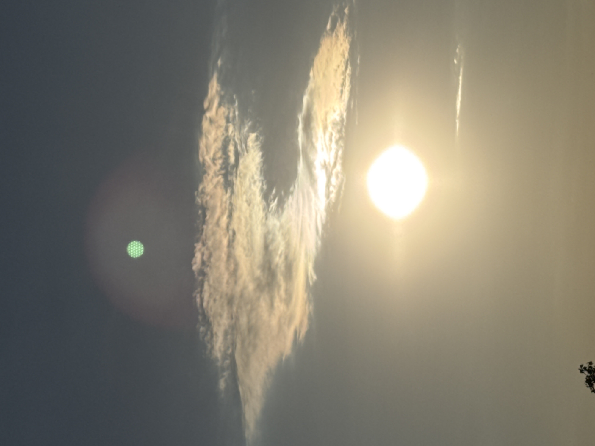 The image depicts a sky with a bright sun low on the horizon, likely during sunrise or sunset. There are several clouds, some illuminated by the sun, casting a warm glow. In the upper part of the sky, there is a distinct green circular object with a patterned texture, possibly a lens flare or an artifact. The lower part of the image shows the silhouette of a tree branch against the bright sky. The scene appears to be an outdoor environment, with no specific city landmarks identifiable, therefore the location in Ruse, Bulgaria cannot be confirmed from the visual information.