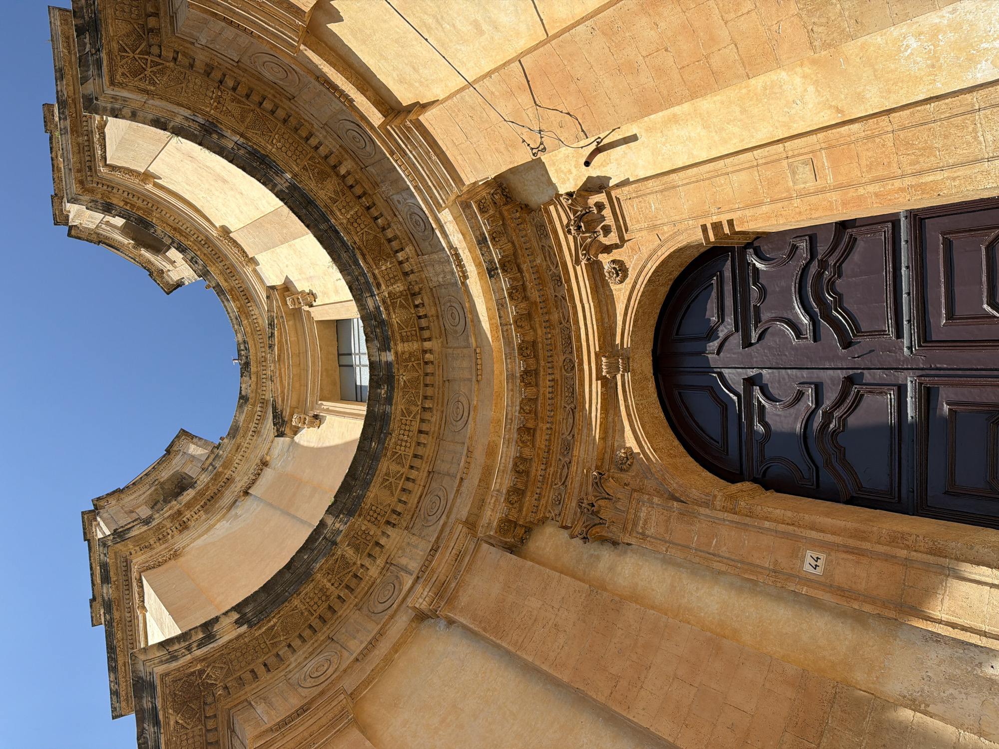 The image shows a low-angle view of architectural details in Noto, Italy. Specifically, the focus is on the facade of a building constructed with light-colored stone, possibly limestone or sandstone, characteristic of Baroque architecture. A prominent arched entrance frames a dark-colored, paneled wooden door. The archway is ornately decorated with carved details and moldings. A window can be seen through the archway. The building has the number "44" on a plaque near the door. The sky is a clear, bright blue, visible through the opening of the arch.