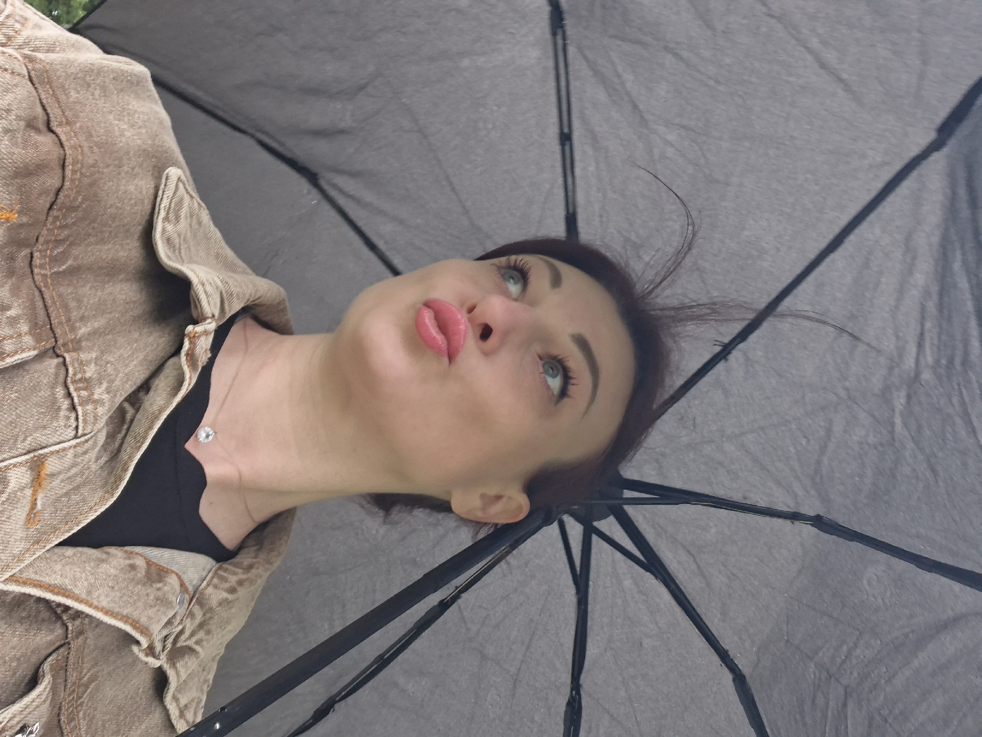 A young woman with auburn hair and blue eyes is holding a dark gray umbrella and looking up. She is wearing a brown denim jacket over a black top. The scene appears to be outdoors, possibly in Забалоцкі сельскі Савет, Belarus, given the user's request.