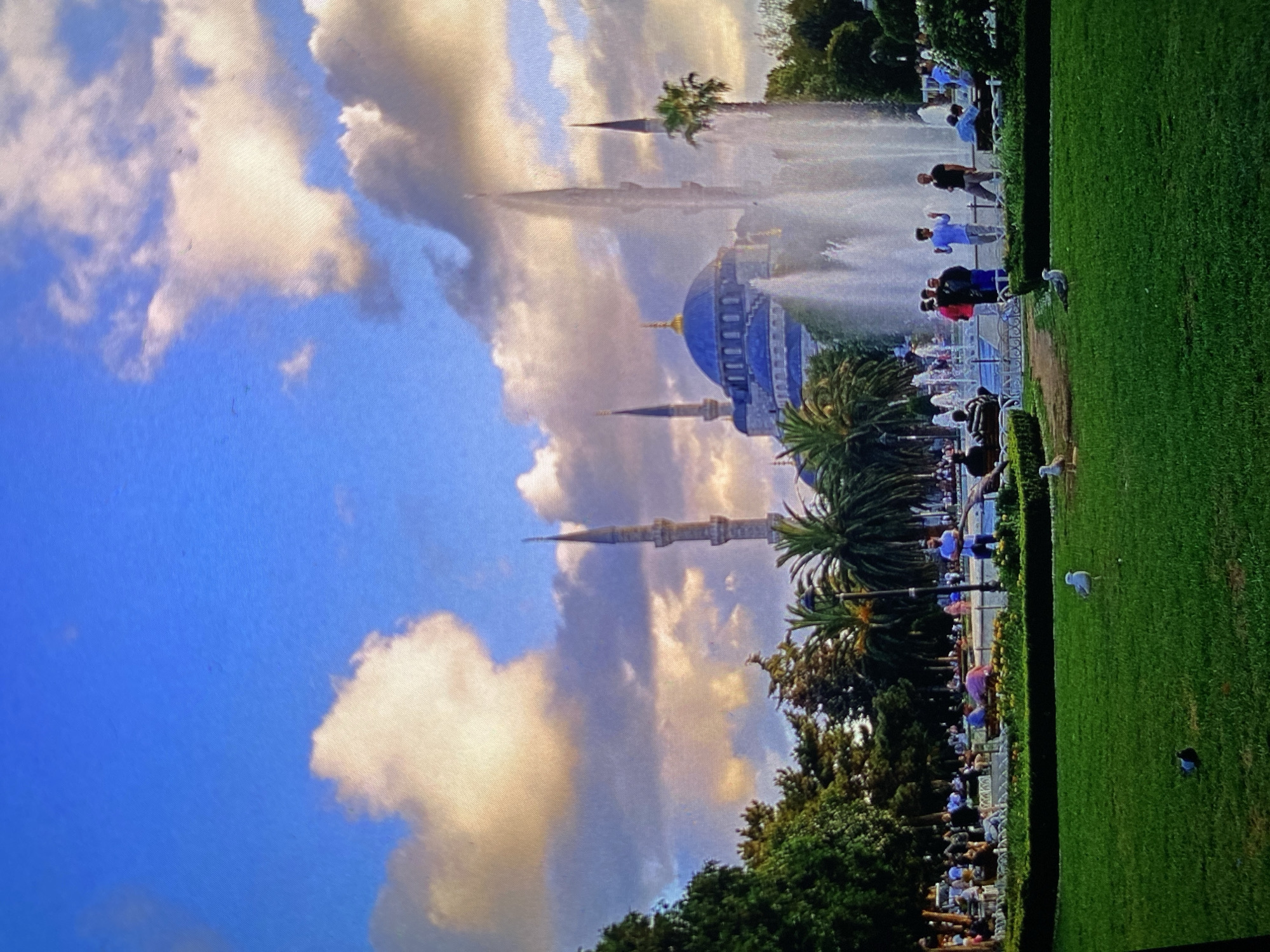 The image, rotated 90 degrees counter-clockwise, displays a scenic view of a grand mosque with a prominent blue dome and multiple slender minarets, unmistakably identified as the Sultan Ahmed Mosque (Blue Mosque) in Istanbul, Turkey, which contradicts the provided location context of Bankya, Bulgaria.
The mosque is framed by a partly cloudy sky, showing patches of clear blue with large, dramatic clouds reflecting warm, golden light, suggesting either late afternoon or early evening. In the foreground and midground, there is an expansive, well-maintained garden area with vibrant green grass and various trees, including several palm trees. A series of large fountains are actively spraying tall streams of water, creating a misty visual effect in front of the mosque.
Numerous people are scattered throughout the garden and around the fountains, appearing to be visitors or tourists enjoying the landmark. They are seen walking, standing, and observing the architecture and water displays. Several birds, resembling seagulls, are visible on the grass. The scene evokes a tranquil and observational atmosphere, capturing a famous historical site under a picturesque sky. No discernible text is visible.
