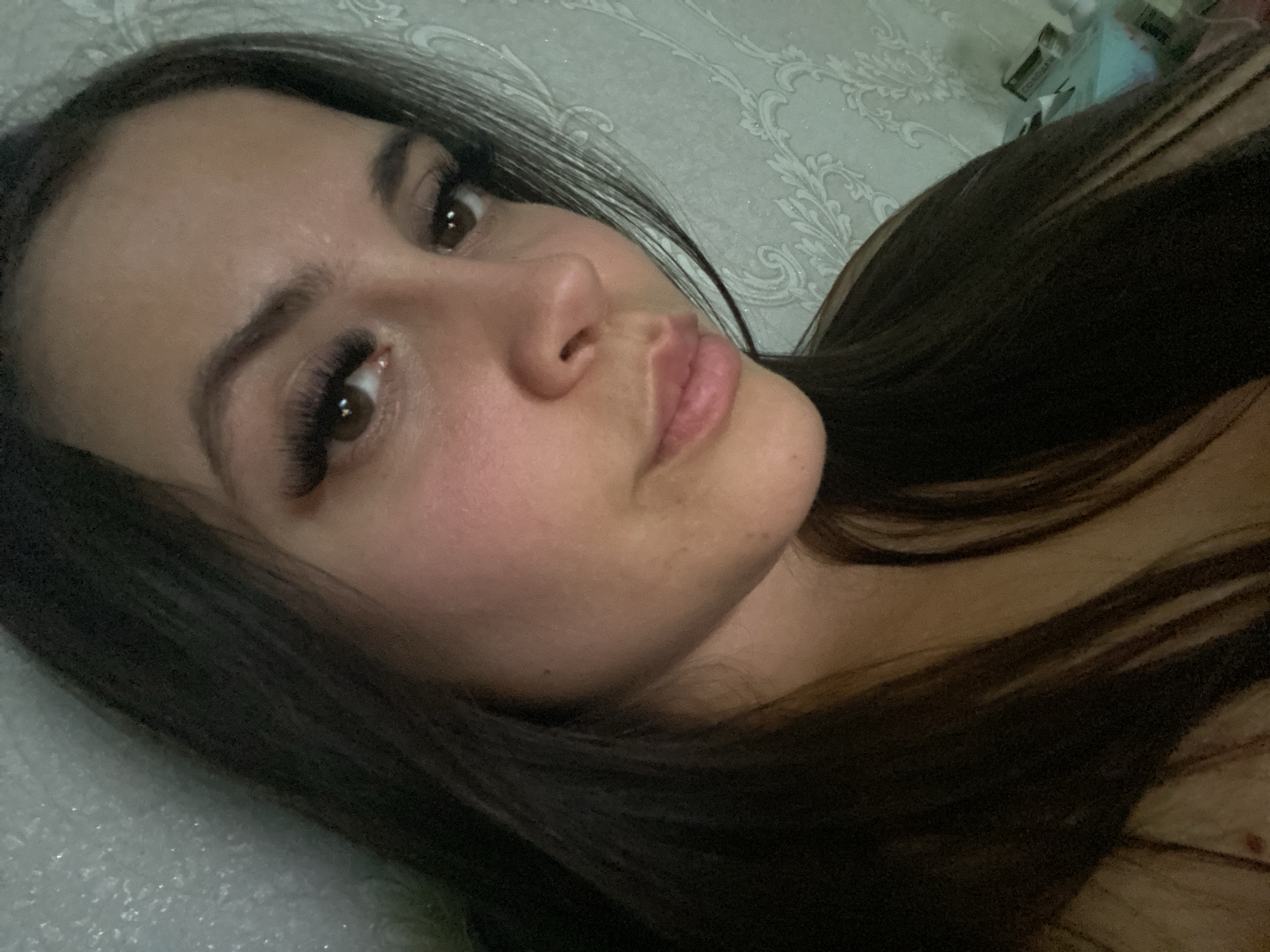 A close-up shot of a young woman's face with dark hair. She has brown eyes and appears to be wearing false eyelashes. Her skin tone is fair, and her lips are slightly parted. The background is blurred, but a patterned wall and some objects possibly related to beauty products are visible, though not clearly distinguishable. The location of Chaikovsky, Russia is not supported by the visual information present.