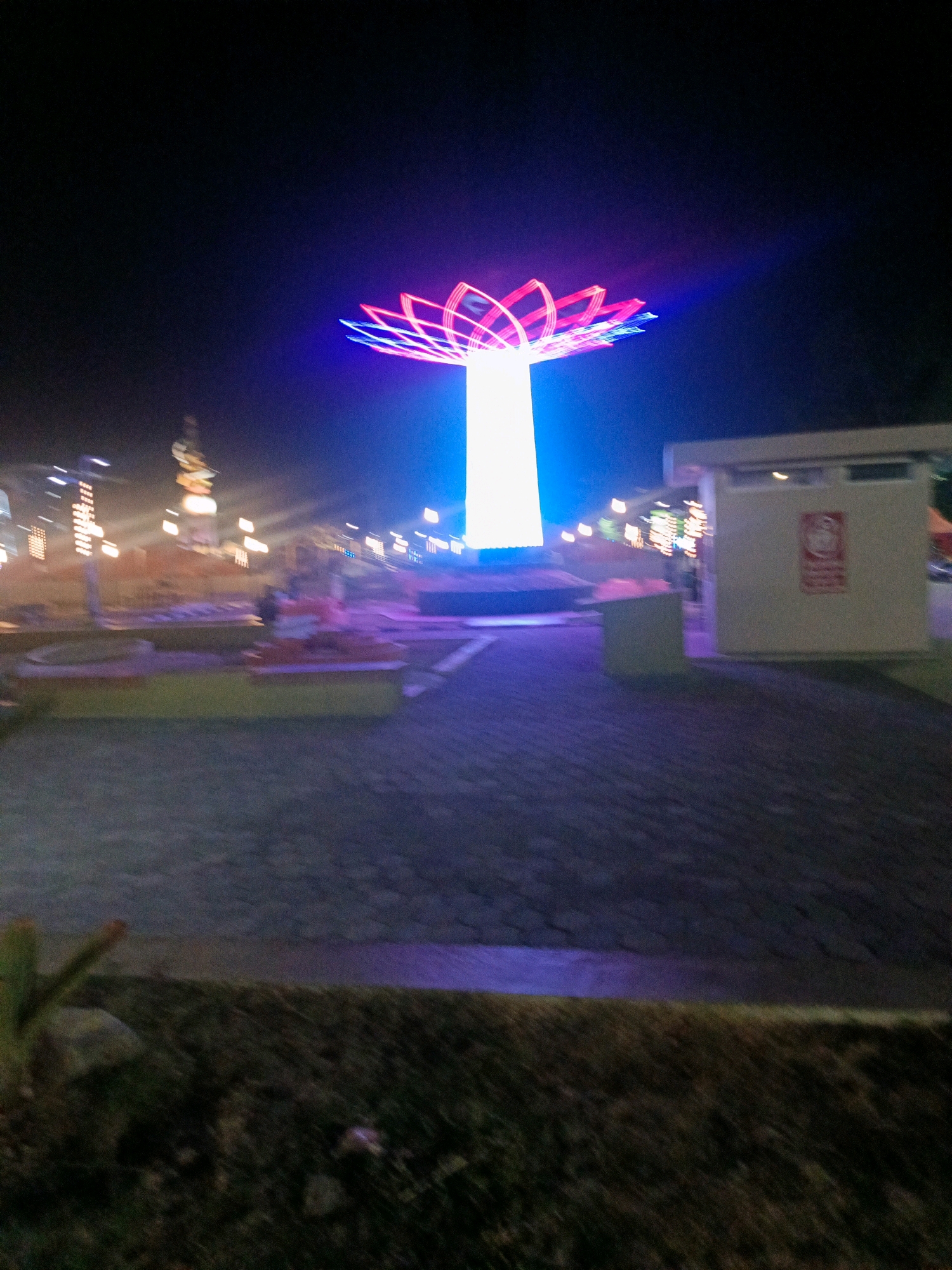 An outdoor nighttime scene features a large, brightly lit structure resembling a lotus flower with pink and blue neon lights at the top, standing tall and white in the center. The surrounding area is paved with dark cobblestones. To the right, a light-colored building with a red emblem is visible. In the background, various illuminated structures, possibly part of a park or entertainment area, are present, including a tall, ornamental tower. The overall ambiance suggests a public space that is active at night. The image is slightly blurred, possibly due to motion or low light conditions.