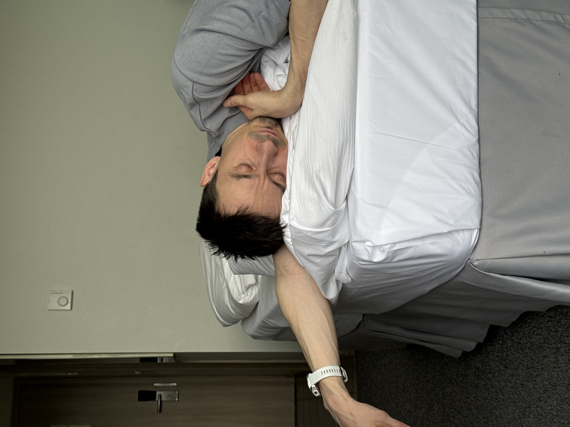 A man is lying in a bed in a room. He is wearing a gray sweatshirt and a white shirt. He has dark hair and is wearing a white watch on his left wrist. The bed has white sheets and a gray bedspread. There is a door and a light switch on the wall. The location is not indicated in the image. The city of Sochi, Russia is not evident from the image.