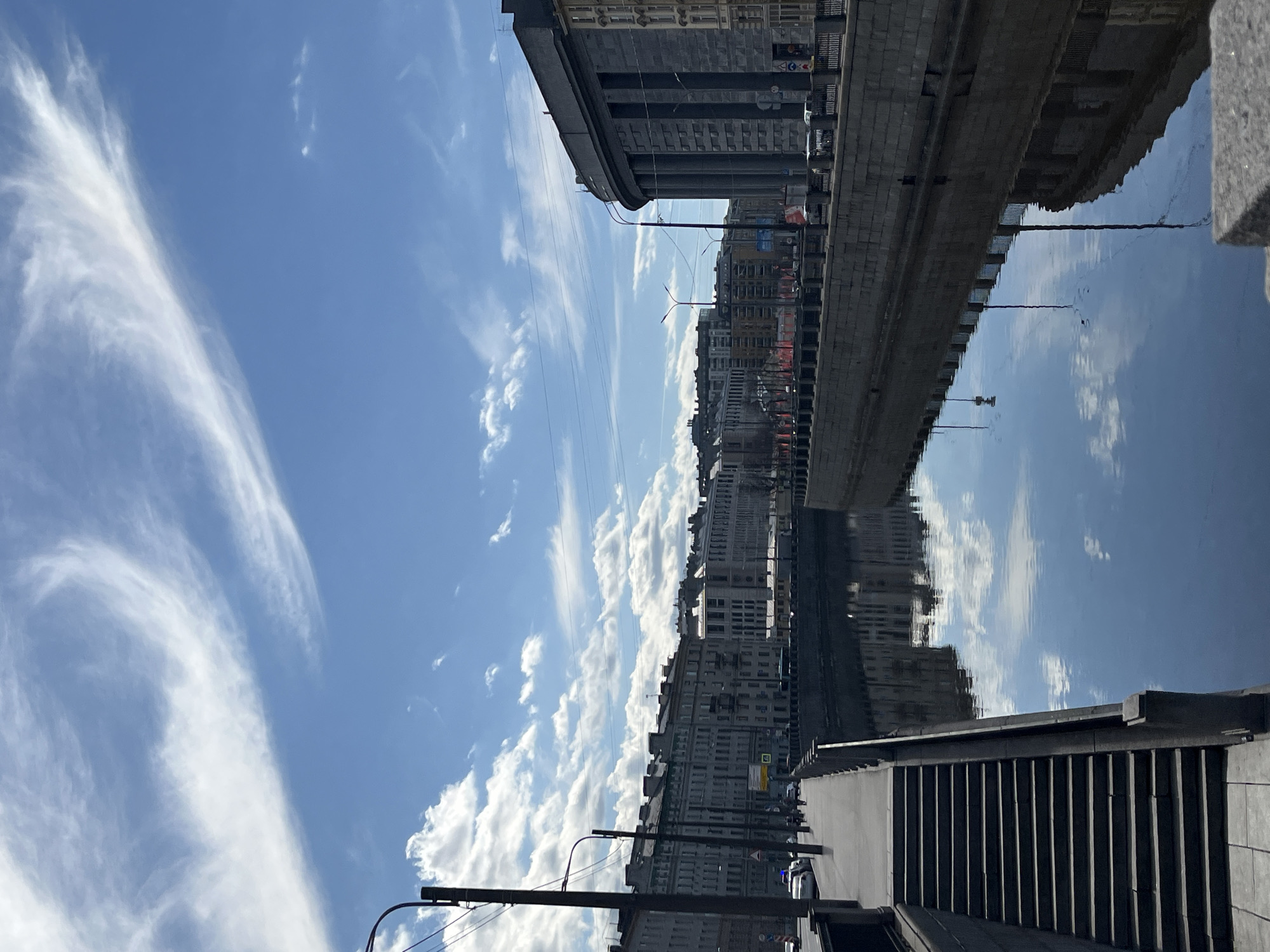 The image captures a bright, sunny daytime scene of a city's architectural landscape bordering a tranquil canal or river. The photograph, oriented vertically, presents a horizontal panorama across the urban environment.
A vast expanse of clear blue sky fills the upper portion, adorned with delicate, wispy white clouds (cirrus) and scattered, puffier cumulus clouds. Power lines or tram wires are visible stretching across the mid-sky.
Along the far bank of the water body, a dense line of multi-story buildings forms the city's backdrop. These structures exhibit classical architectural styles, featuring numerous windows, intricate facades, and a predominantly grey or beige color palette. A notable detail on one of these buildings is a clear "M" symbol on a white sign with a red border, commonly associated with metro or subway stations in certain regions. Other indistinct signs and distant streetlights can also be seen.
A robust, dark-colored embankment wall or bridge structure runs along the edge of the water, separating the city buildings from the canal. The water below is remarkably calm and still, acting as a mirror to the sky above. It perfectly reflects the blue sky, the white clouds, and the inverted images of the buildings and the embankment, creating a striking symmetrical effect.
In the foreground, on the near bank, a concrete or stone stairway with prominent vertical risers descends towards the water. Parts of streetlights and railings are also visible, along with a few cars on a street running parallel to the canal. The scene evokes a sense of urban tranquility on a clear day, with the vivid reflections adding depth and visual interest. No specific human activity or event is explicitly depicted.