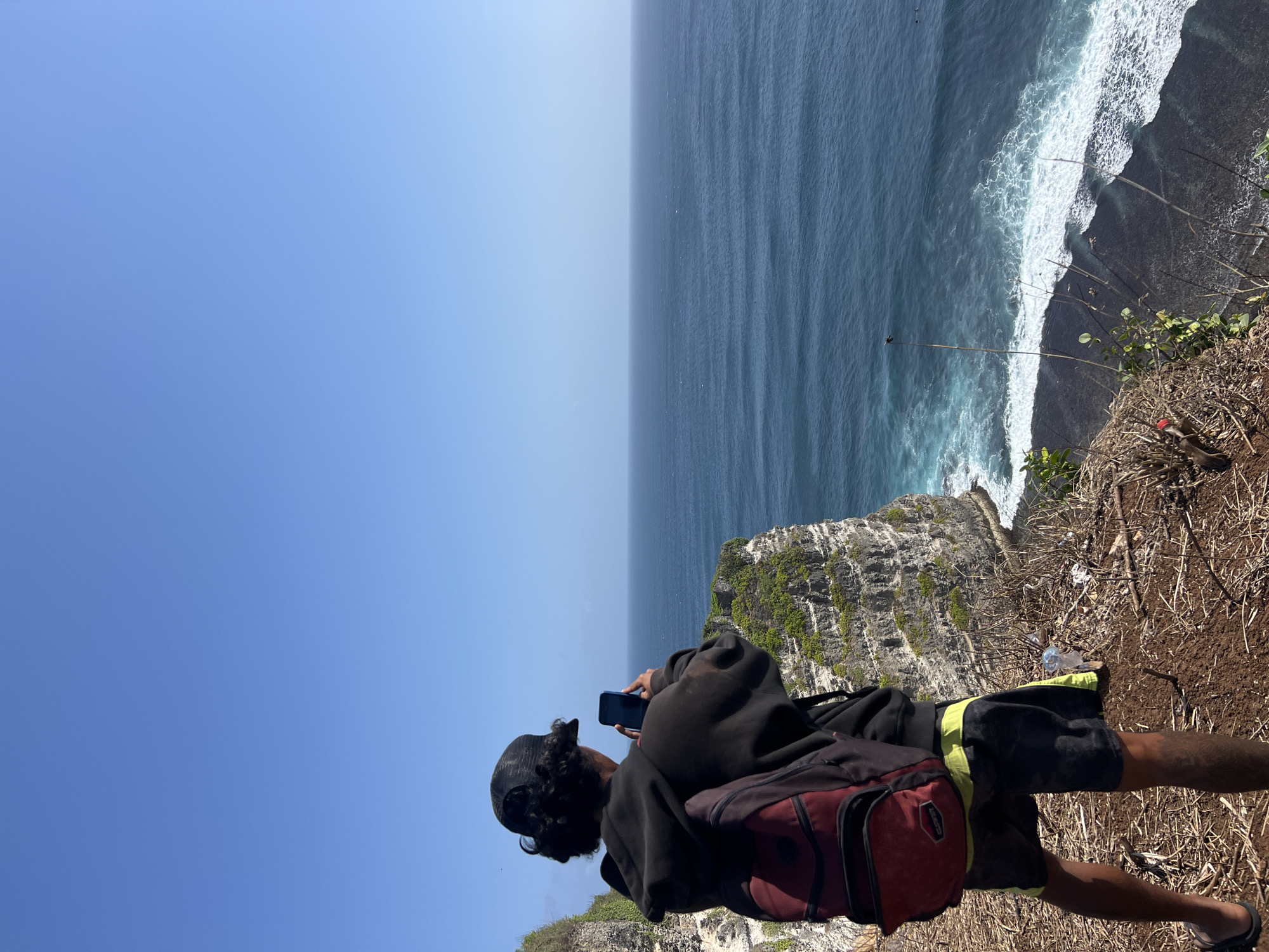 A person wearing a black cap, a black hoodie, and shorts stands on the edge of a cliff, holding a smartphone to take a picture of the ocean. The person has a red and black backpack on their back. The cliff is covered with green vegetation and has a layered rock formation. The ocean is a deep blue with white waves crashing on a dark shore below. The sky is clear and blue. The location is identified as Kuta Selatan, Indonesia.
