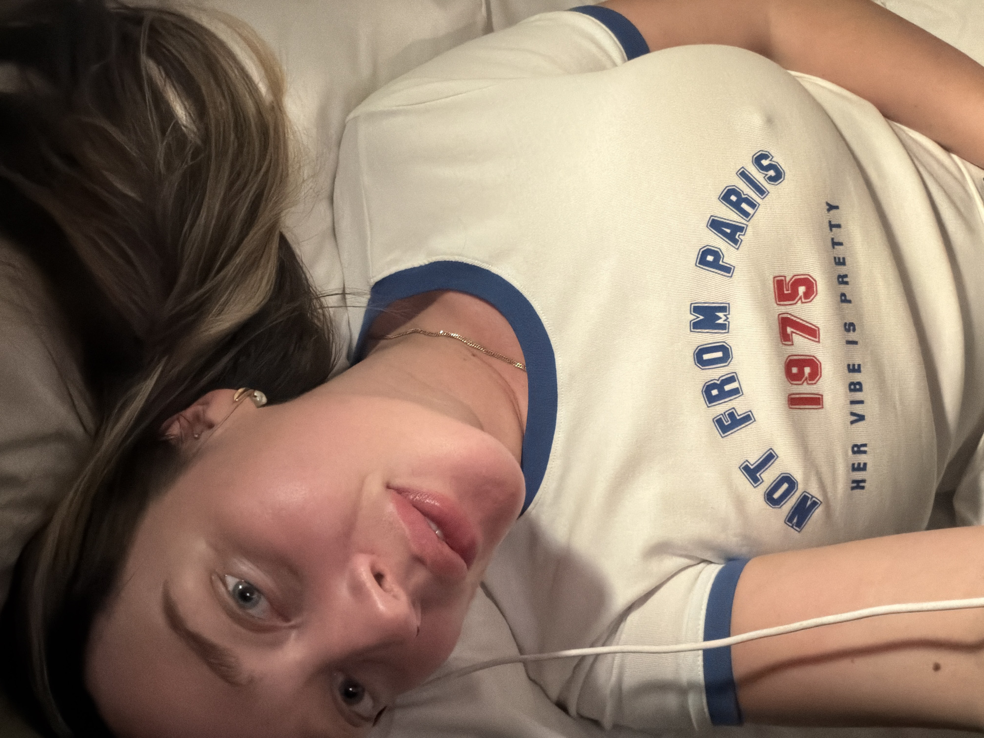 A woman lies on a white surface. She is wearing a white t-shirt with blue trim and blue lettering that reads "Not From Paris," "1975," and "Her Vibe is Pretty." She has light skin, light eyes, and brown hair. She is wearing a gold chain necklace and a gold earring. A white cord lies across her right arm. The location is not specified in the image, so no information about Moscow, Russia can be given.