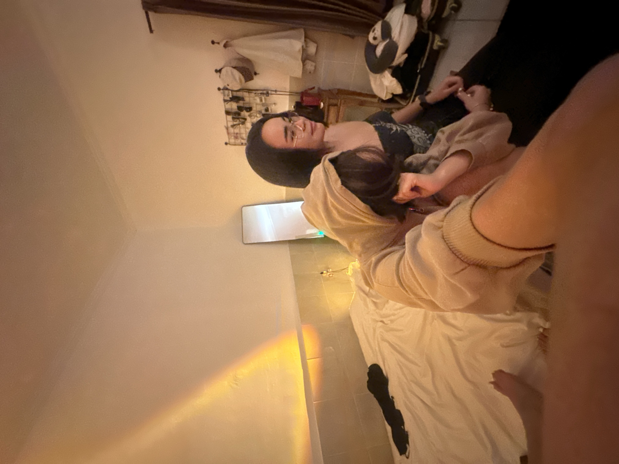 A rotated indoor shot depicts two individuals in an indeterminate space. One individual, wearing glasses and a dark top, is reclined on a dark surface. The other individual is wearing a tan hooded sweatshirt. A bed covered with a white sheet, on which lies a dark object, is visible. Light reflecting from a wall suggests a possible window or light source. A rack with various items and a piece of furniture are visible in the background. The image may have been taken in North Kuta, Indonesia, although that is unconfirmed from the visual data.