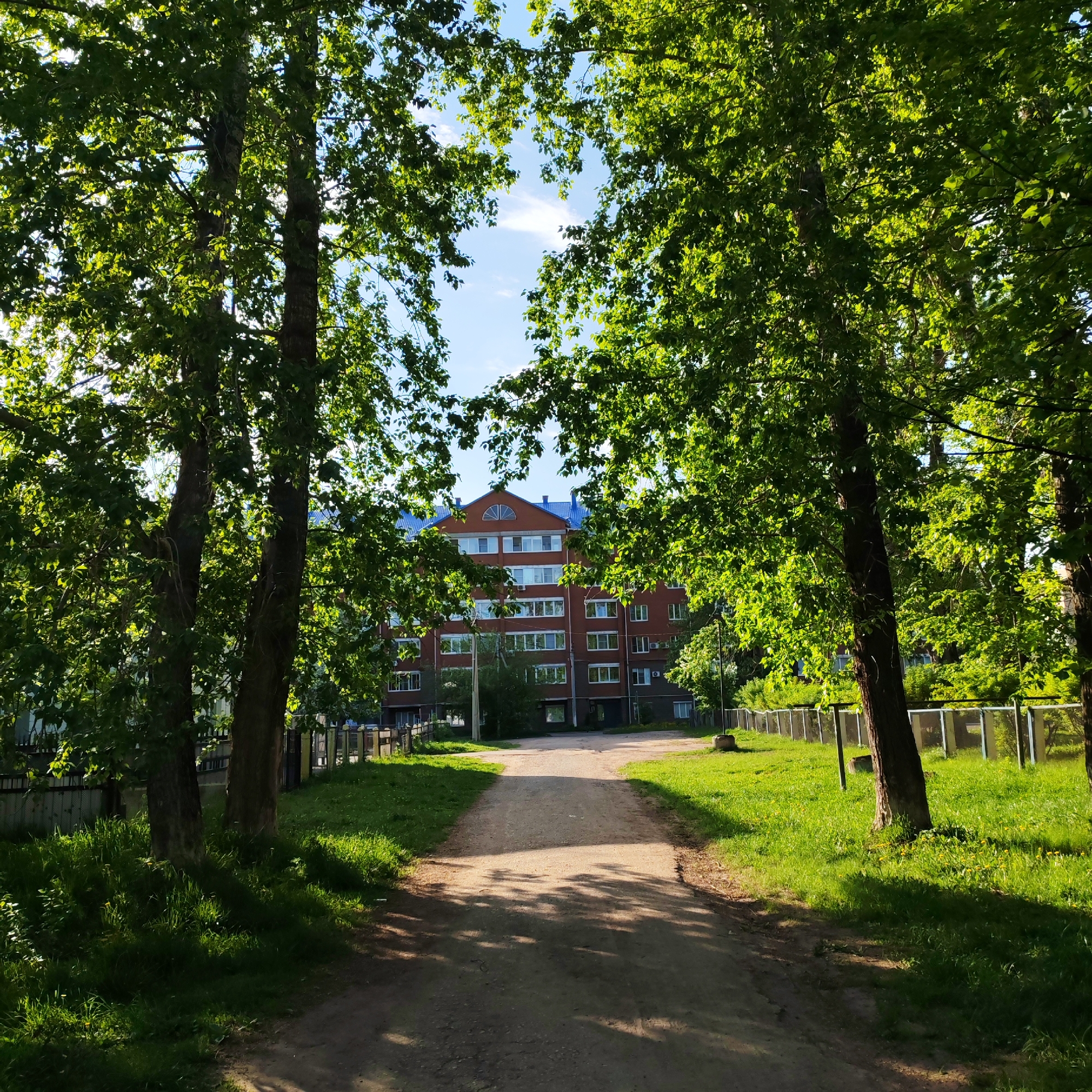 The image depicts a dirt road leading toward a brick building with a blue roof in Chaikovsky, Russia. The road is flanked by green grass and trees that partially obscure the building. A fence runs along the right side of the road. The sky is visible through the trees, indicating a clear day. No people are visible.