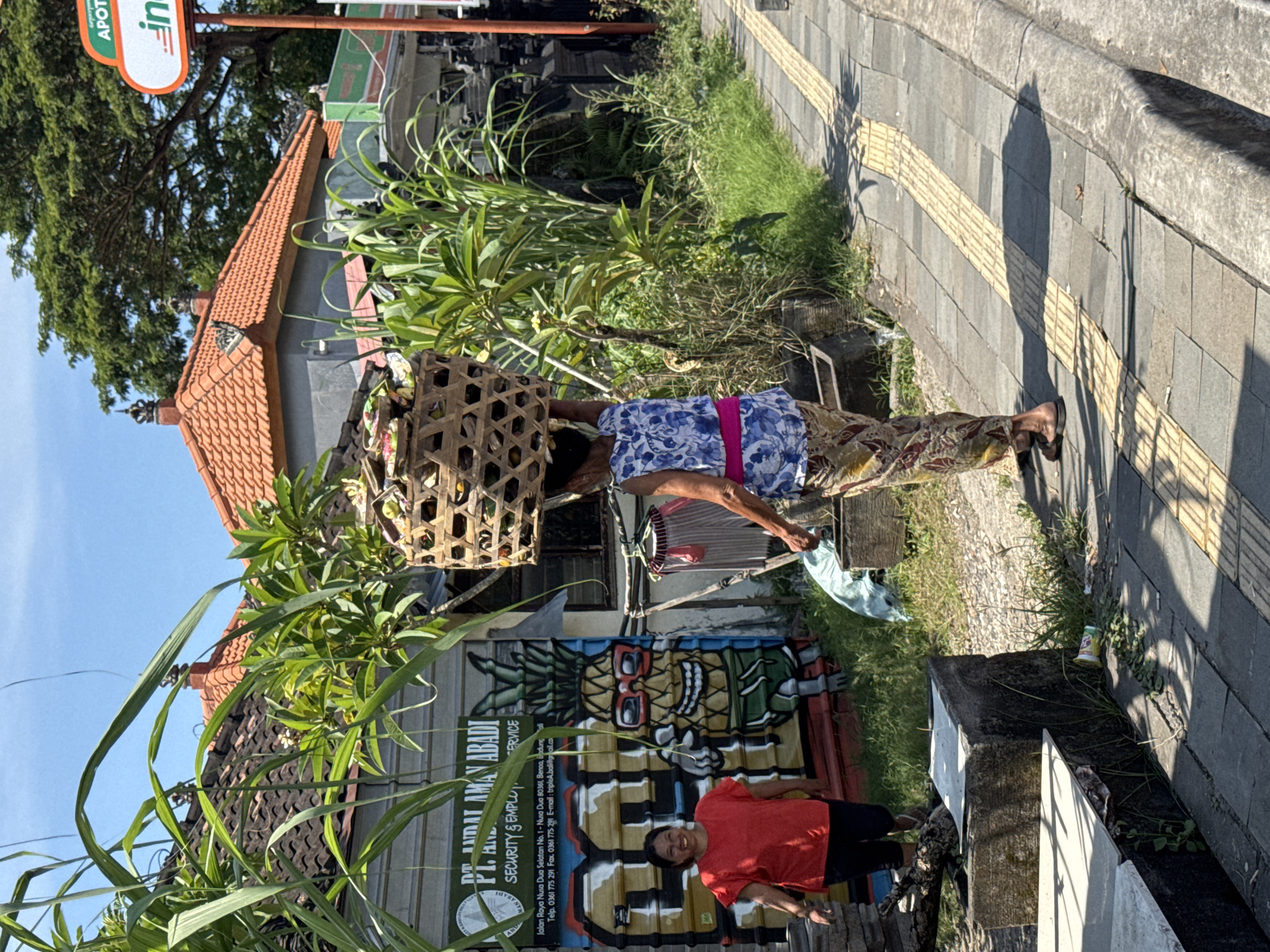 Here's a description of the image:
The image is an outdoor shot featuring a scene in Nusa Dua, Indonesia. A woman wearing a blue patterned top, brown patterned pants, and a pink sash is carrying a large, woven basket on her head. She is walking on a tiled sidewalk along the street.
In the background, there's a building with a reddish-orange tiled roof. A sign for "PT. ADAT AMAN ABADI SECURITY & EMPLU. ERVICE" is visible, with a pineapple-themed graffiti mural on a corrugated metal wall behind it. A person in a red shirt is standing in front of the mural. There are also various plants and greenery in the scene. A sign for "APOTEK" can be seen in the background.